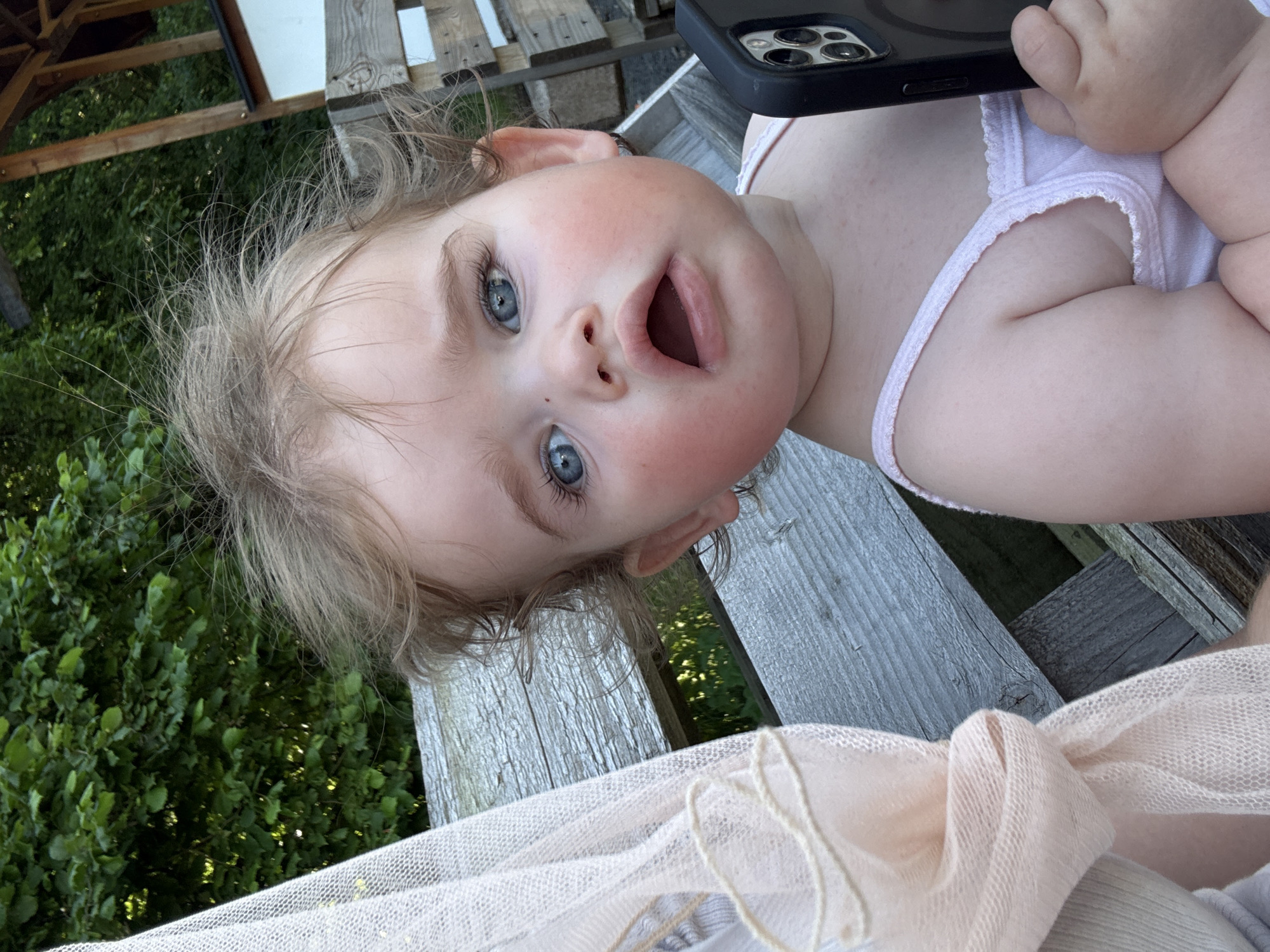 A young child with blonde, messy hair and bright blue eyes is looking directly at the camera with an open mouth. The child is wearing a light pink top with a ruffled edge. To the left of the child, a light pink, sheer fabric is draped. The child is holding a black smartphone in their right hand. The background consists of a wooden bench and greenery. The scene appears to be outdoors, possibly in a park or backyard setting in Beltershain, Germany.