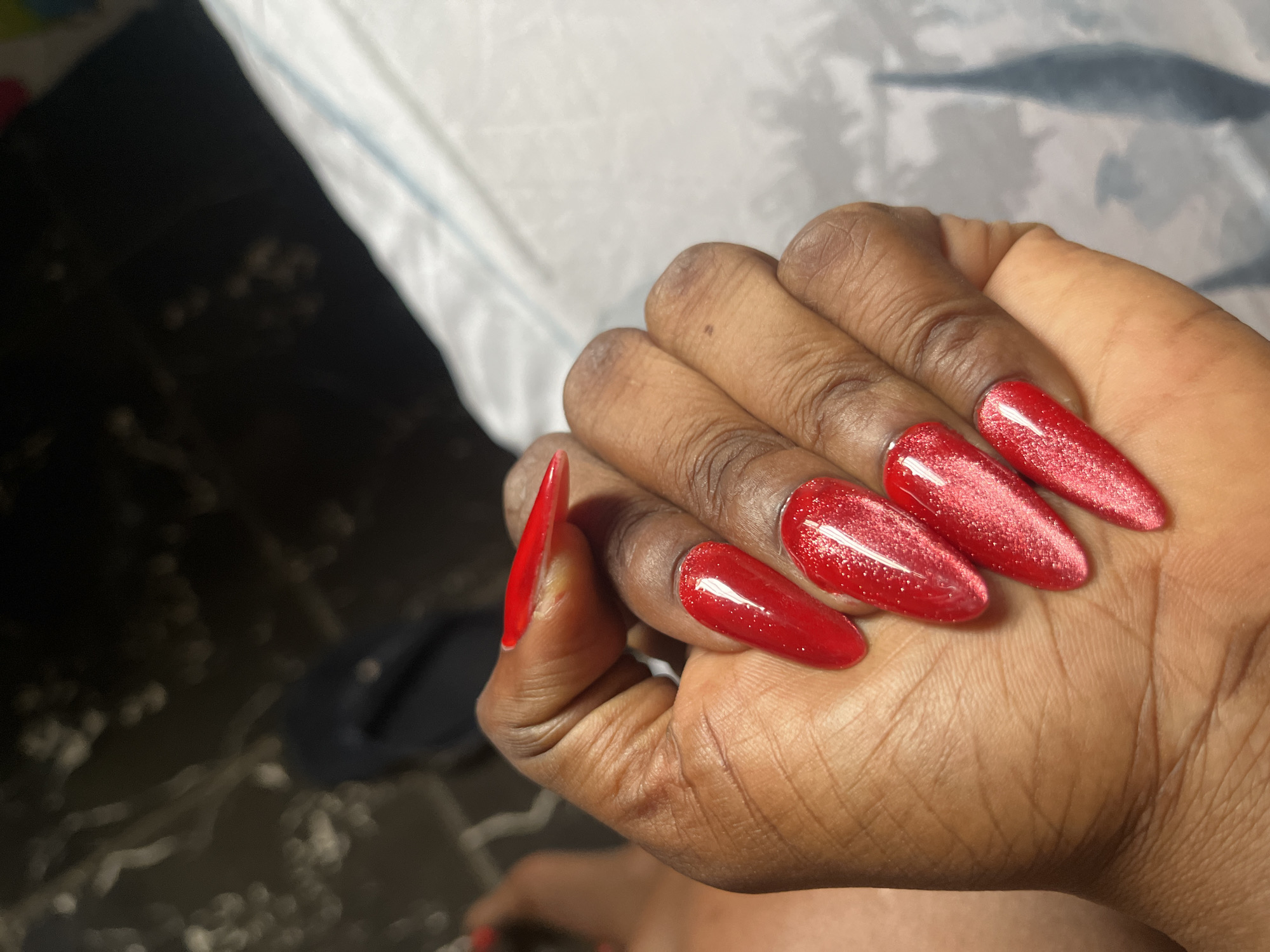 A close-up, high-angle shot focuses on a person's hand with long, almond-shaped, bright red glitter-polished fingernails. The hand is positioned in the foreground, with the thumb and index finger prominently visible, along with three other nails. The skin tone is dark. In the background, out of focus, is a dark surface with some indistinct lighter shapes and a white, textured surface. A dark blue flip-flop is visible in the far left background. The overall lighting is dim. No specific location in Jos, Nigeria, can be identified from this image.