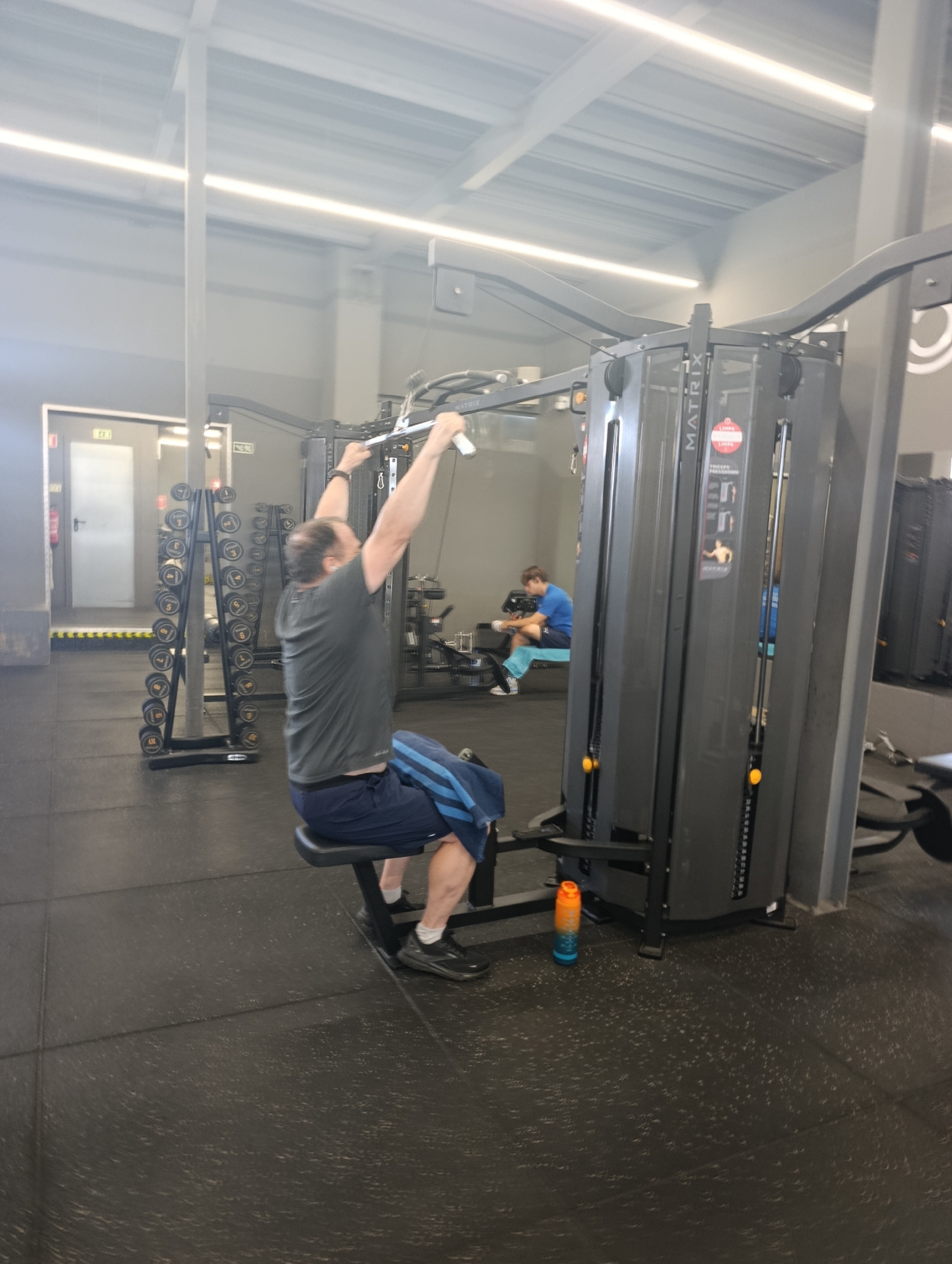 The image depicts an indoor gym environment, likely in Setúbal, Portugal, given the presence of the word "LIMPA" (Portuguese for "clean") on a sticker on the gym equipment. In the foreground, a man with short brown hair is seated on a black exercise bench, performing a lat pulldown exercise on a grey Matrix cable machine. He is wearing a dark grey t-shirt and dark blue athletic shorts with light blue stripes. His arms are extended upwards, gripping a silver bar attached to the cable. To the left of the man, a rack of dumbbells is visible, with weights ranging from what appears to be 10 to 40 kilograms. In the background, another person is seated on a bench, wearing a blue t-shirt and white shorts, and looking down at something in their hands. The gym floor is black rubber matting, and the walls are a neutral grey. Fluorescent lighting fixtures are visible on the ceiling. A bright orange and blue water bottle rests on the floor near the cable machine. The overall scene suggests a typical fitness facility.