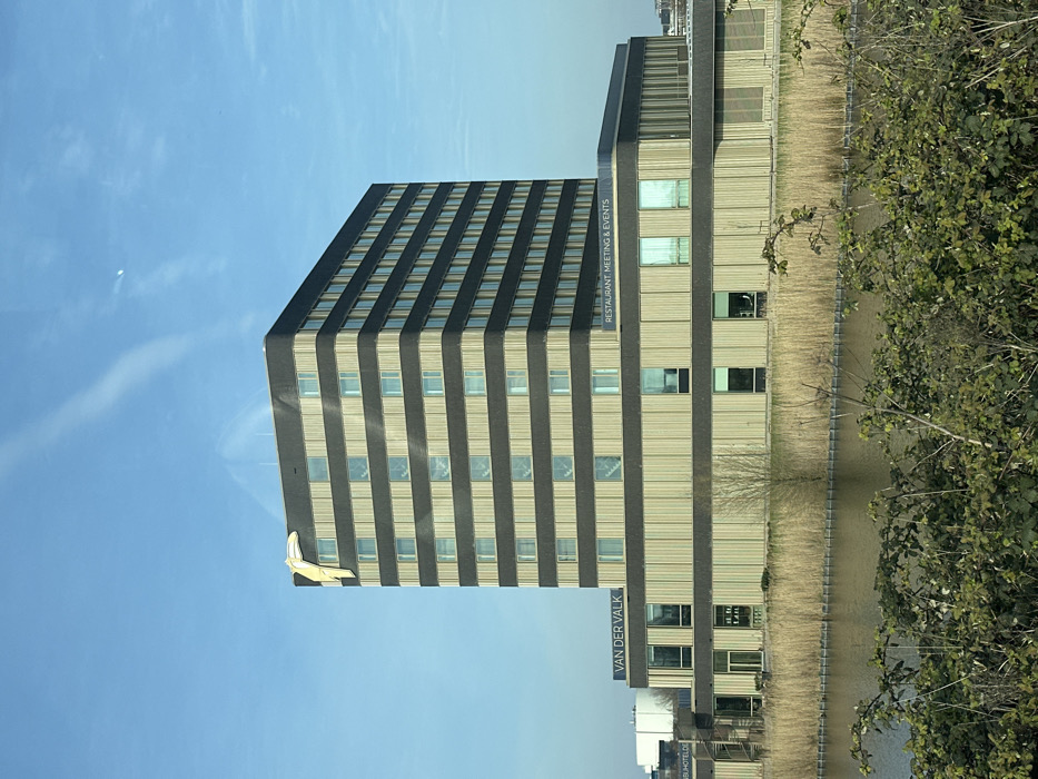 The image displays the Van der Valk hotel in Den Hoorn, Netherlands, under a clear blue sky. The prominent structure is a multi-story building featuring a dark-paneled tower with ten visible floors, each horizontally striped with lighter, vertically-slatted sections and multiple reflective rectangular windows. A large, stylized yellow bird figure is affixed to the upper left corner of the tower. Below the tower, a two-story base extends horizontally, bearing the text "VAN DER VALK" on its left section and "RESTAURANT. MEETING & EVENTS" on its right. This base also incorporates light-colored vertical slats and rectangular windows. In the midground, a body of still water, likely a canal, stretches across the frame, its far bank densely lined with dry, tall reeds or grasses. The immediate foreground contains green, leafy shrubbery, partially obscuring the water. No individuals are clearly visible within the scene.
