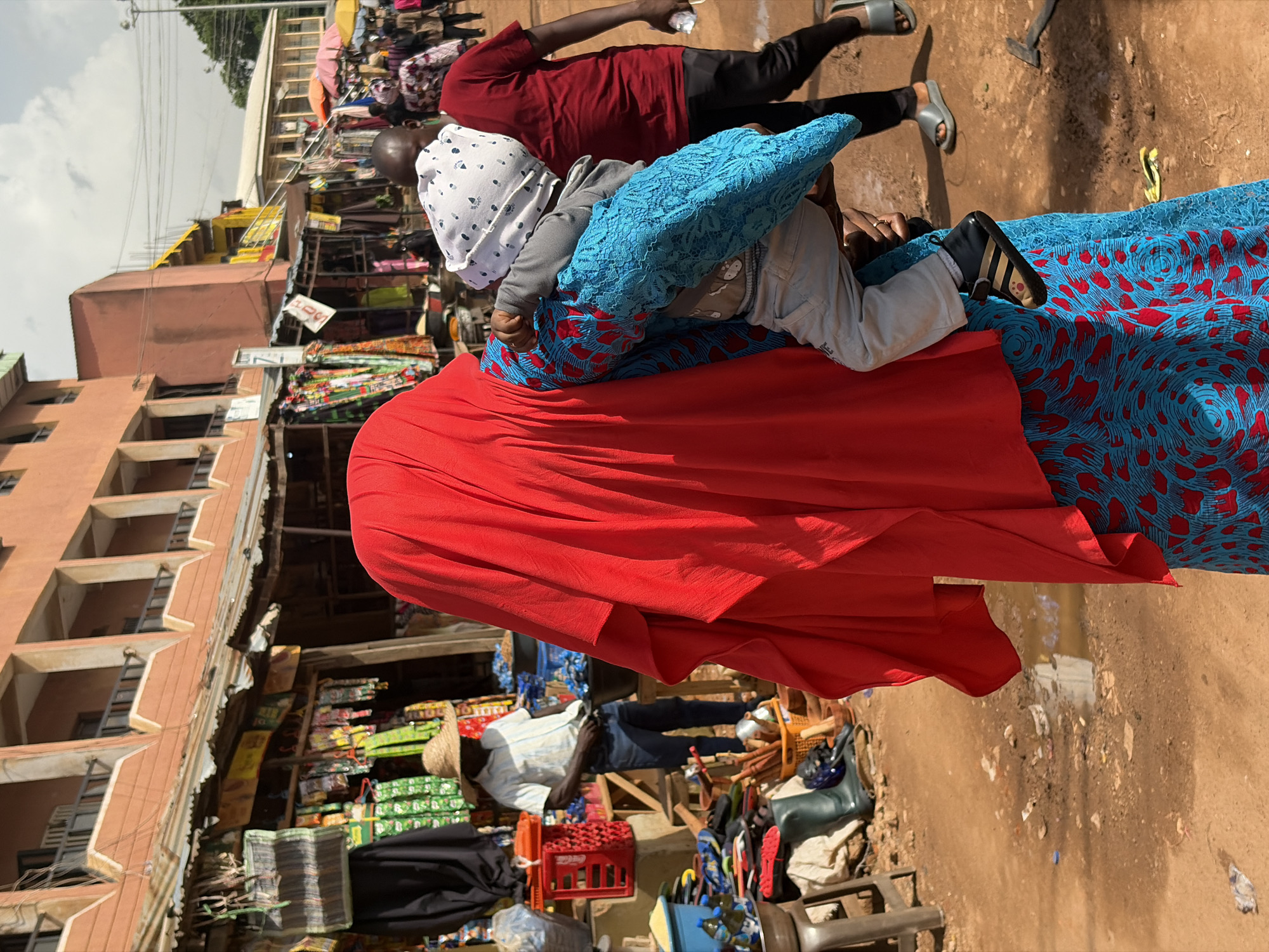 In a bustling outdoor market in Jos, Nigeria, a woman is carrying a child wrapped in a blue and red patterned fabric. The child is wearing a white patterned cloth on its head and grey long-sleeved shirt and light-colored pants. The woman is wearing a red garment that covers most of her body, and a blue and red patterned dress is visible beneath it.
In the background, other people are visible, including a man in a red shirt and dark pants, and another man wearing a straw hat, a light-colored striped shirt, and dark pants. He is looking at his phone. Various stalls with goods are set up in the market, displaying colorful textiles, crates of items, and other merchandise.
The ground is a mix of dirt and some puddles, suggesting recent rain or a damp environment. The buildings in the background appear to be multi-story structures, typical of urban settings. The sky is overcast, indicating cloudy weather. The scene captures a slice of daily life in a Nigerian market, with people engaged in their activities, including shopping, carrying children, and possibly tending to their stalls. There are no clearly legible signs with text visible in the image.