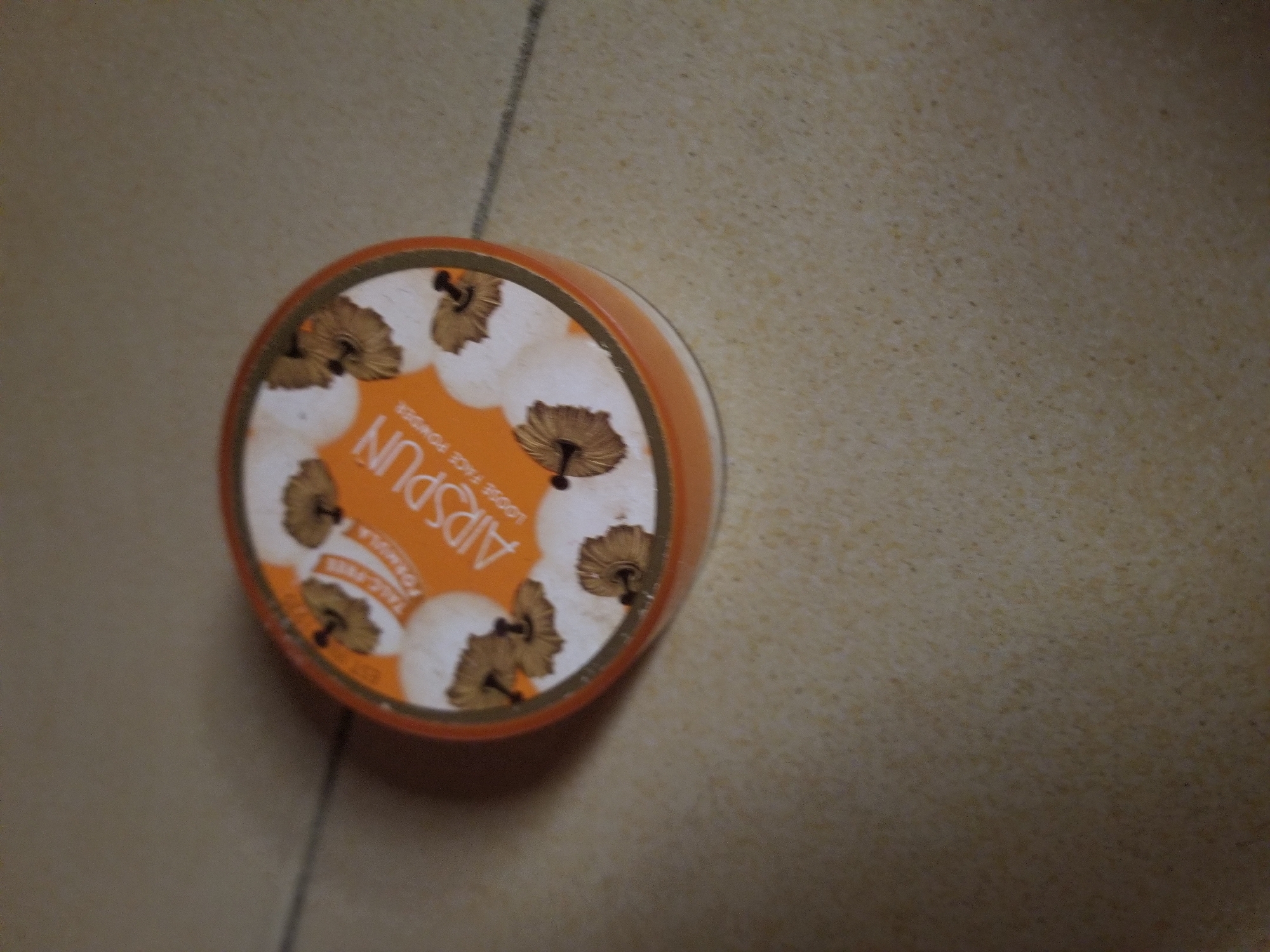 The image shows a close-up, slightly overhead view of an orange and white round container of Coty Airspun Loose Face Powder. The container is on a light-colored, speckled surface, possibly a countertop or floor. The lid of the container is visible, featuring the brand name "AIRSPUN" in white lettering against an orange background. The lid also has a decorative pattern of white circles with brown, fan-shaped designs inside. The powder inside is not visible. There are no people or other objects present in the immediate vicinity. The lighting appears to be indoor, casting a slight shadow. The location of Jauro Yinu, Nigeria is not depicted or implied in the image.