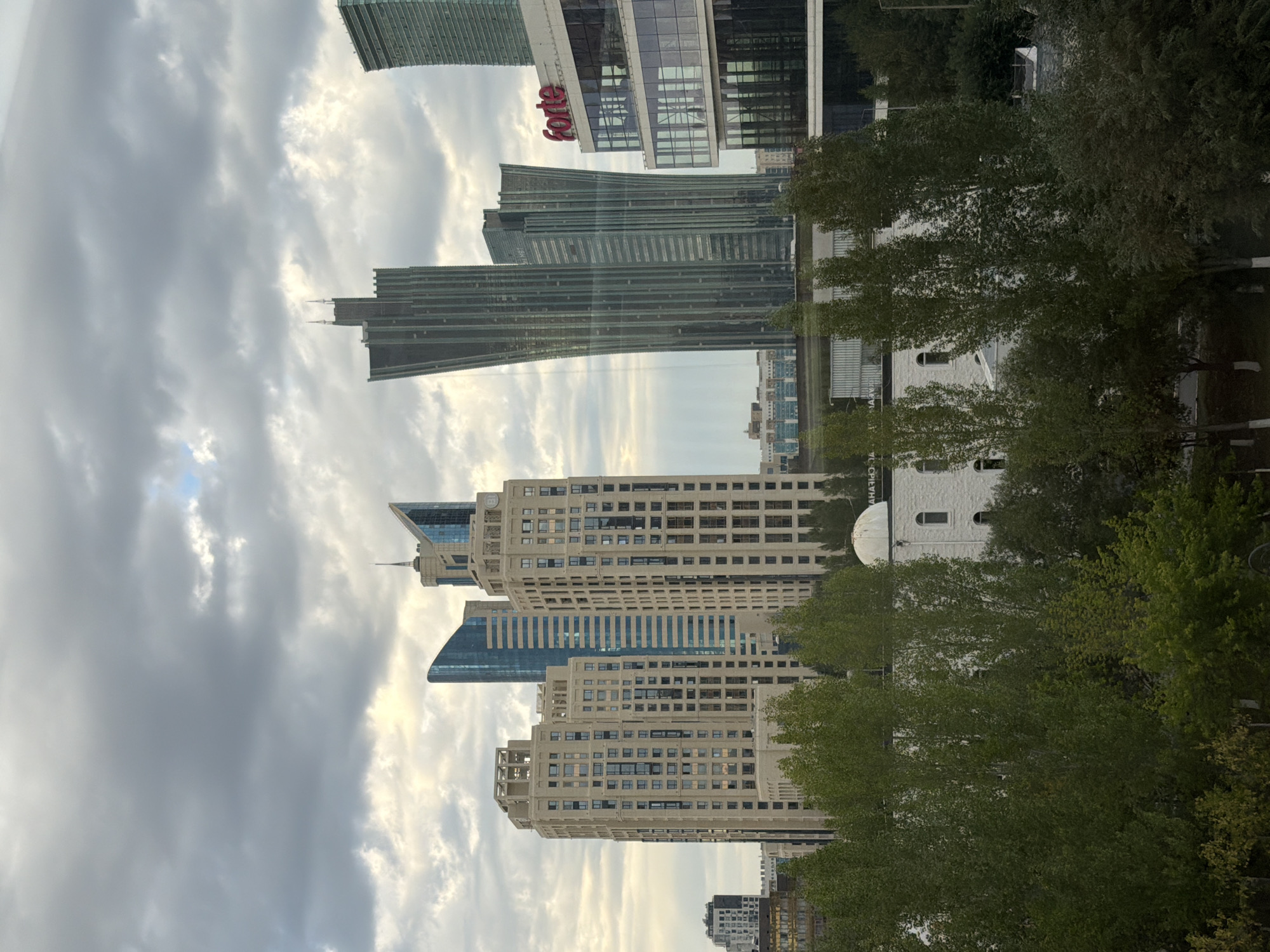 The image depicts a modern urban landscape in Astana, Kazakhstan, under a cloudy sky. The foreground is dominated by a dense canopy of green trees, with varying shades of green indicating different species or leaf maturity. Beneath the trees, hints of the urban environment emerge, including the top of a white building with a domed roof and arched windows.
In the mid-ground, a cluster of tall, contemporary buildings rises. To the left, several beige-colored residential towers with numerous windows stand, some featuring visible balconies. Behind them, a sleek blue skyscraper with a curved facade adds a contrasting architectural element. Further to the right, an imposing dark green skyscraper with a distinctive angular design pierces the sky. Adjacent to this is a modern glass-fronted building with a prominent red "forte" sign on its upper facade.
In the background, more buildings are visible, some appearing as lower-rise structures and others as distant high-rises, suggesting a sprawling city. The overall atmosphere is one of urban development and modern architecture, with the overcast sky creating a somewhat subdued lighting condition. No people or discernible activities are visible in the image.