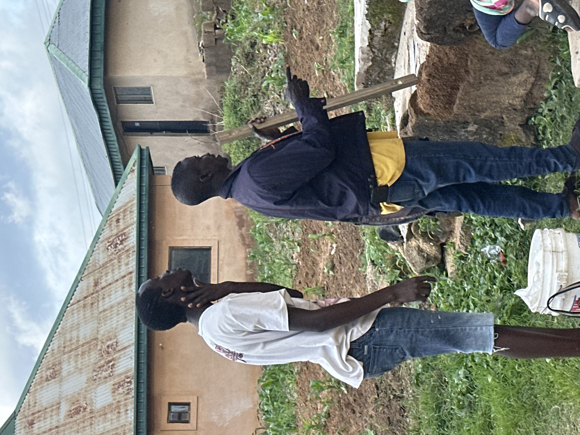 The image shows two young Black men in Gwong, Nigeria. The man in the foreground is wearing a white t-shirt and denim shorts. He is looking up and to his left, with his hands clasped around his neck. He appears to be experiencing distress or discomfort. Behind him, another man is wearing a navy jacket over a yellow shirt and denim jeans. He is holding a long, thin wooden object horizontally in front of him and appears to be pointing his left index finger upwards, possibly giving instruction or explanation.
The scene is outdoors, with a dirt and grass-covered ground. Two buildings with corrugated metal roofs are visible in the background. The sky is overcast. The overall impression is a casual, possibly rural setting. There are no visible signs of text in the image.