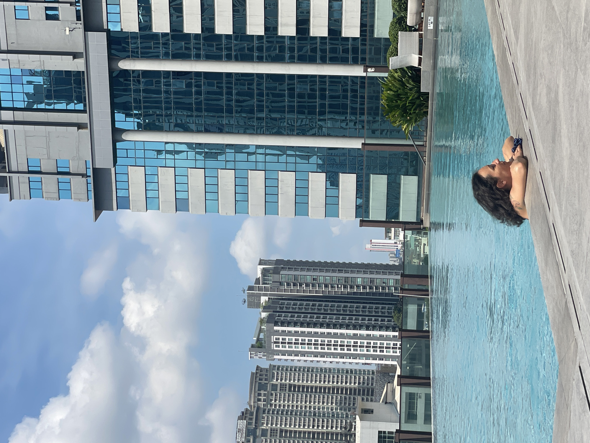 A vertical outdoor shot features a person in a rooftop swimming pool, with a cityscape of Bangkok, Thailand in the background. The foreground includes a section of the pool with clear blue water and the concrete edge of the pool. A woman with curly hair is visible in the water, leaning against the edge of the pool. Her eyes are facing towards the upper right. In the midground, a variety of tall buildings are visible. The buildings vary in height and design, some with reflective glass windows. A sky is partially visible behind the buildings with scattered clouds.