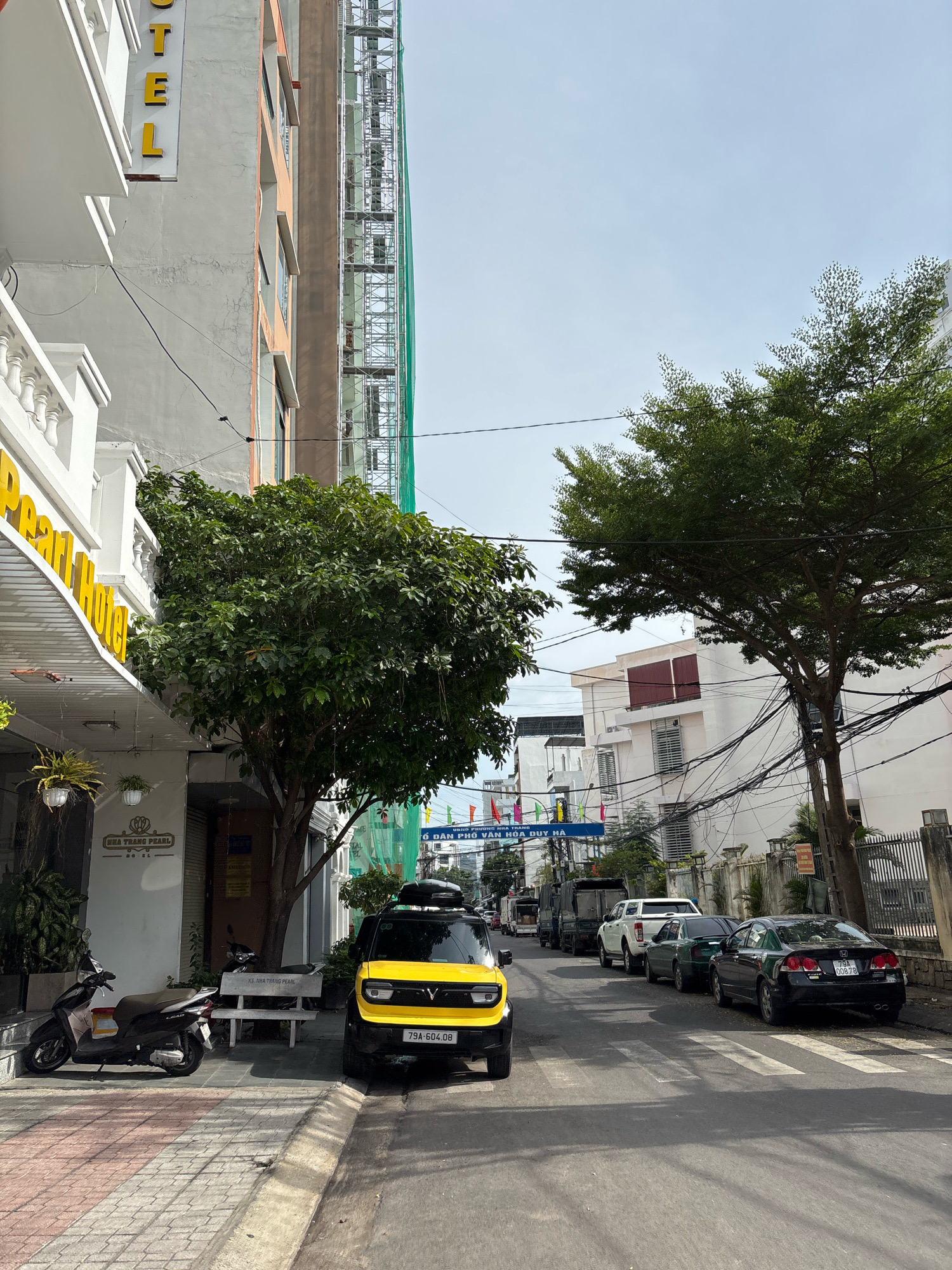 The image depicts a street scene in Nha Trang, Vietnam, likely in the vicinity of Nha Trang Pearl Hotel, as indicated by signage. The perspective is from a low angle, looking down a street lined with buildings. On the left, a white building with yellow signage that reads "HOTEL" and part of "Pearl Hotel" is visible, with a white balcony railing. Adjacent to it is a green tree with dense foliage. In front of the hotel, a dark-colored scooter is parked on a sidewalk paved with brick-patterned tiles. A white bench with "KS. NHATRANG PEARL" is also present.
Further down the street, a bright yellow compact car with a roof-mounted cargo box is parked. Its license plate reads "79A-604.08". Several other vehicles are parked along the street, including a black sedan with license plate "79A 00878", a dark green sedan, and a white pickup truck. A large truck is also visible in the background. The street is paved with asphalt and features a pedestrian crossing.
Buildings on the right side of the street are varied in architecture, with some appearing to be white with modern windows and others with older facades. A construction site, covered with green netting, is visible behind one of the buildings. Overhead power lines are strung across the street, a common feature in many urban areas in Vietnam. Colorful flags are displayed on poles in the distance, adding a touch of vibrancy to the scene. The sky is clear and light blue, suggesting a sunny day. The overall environment is that of a bustling urban street with a mix of residential and commercial establishments, alongside everyday traffic.