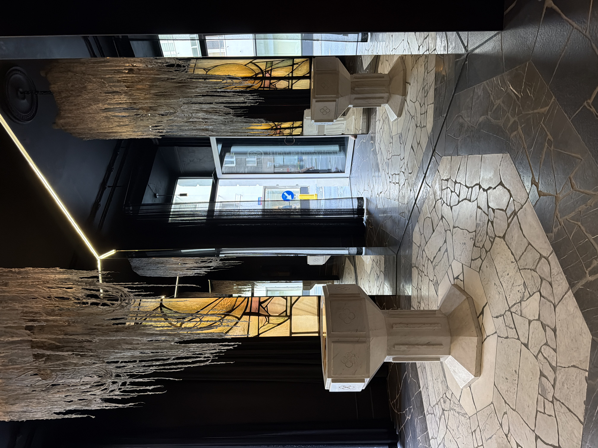 The image depicts an indoor space characterized by an eclectic design, featuring two large, decorative structures resembling baptismal fonts or pedestals, a pair of unique chandeliers, and extensive use of mirrors. The location is an interior space in the city of Unknown, Unknown.
In the foreground and slightly to the left, a light-colored, octagonal pedestal or font stands centrally. It is made of a stone-like material, possibly marble or concrete, and features carved quatrefoil designs on its upper facets. The bowl is shallow and reflects a yellow light from above.
Above this pedestal, suspended from the dark ceiling, is a large, white chandelier composed of numerous long, thin, textured strands or fibers, resembling a waterfall or icicles. Its reflection is visible in the tall, vertical mirrors that dominate the right side of the image.
The ceiling is dark, possibly black or dark grey, and features a linear LED light strip running along a corner, providing a warm yellow glow. A dark ceiling rose is visible where the chandelier chain attaches.
Behind the left pedestal, dark curtains frame what appears to be a stained-glass panel with abstract geometric patterns in shades of yellow, amber, and some darker hues. This panel is also reflected in the large mirror directly to its right.
The right side of the image is largely defined by a large, vertical mirror, creating a symmetrical reflection of the left half of the room. A second identical pedestal is visible further back in the reflection, positioned similarly to the primary one. Another chandelier reflection is also prominent.
Beyond the mirrored wall, an open glass door reveals an outdoor street scene. A blue circular street sign with a white arrow pointing left is visible on a yellow pole. Buildings and other street elements are visible outside. The reflection of this street view is also visible in the mirror.
The floor is a mosaic or terrazzo pattern composed of irregularly shaped tiles. In the center, beneath the primary pedestal, there is a lighter-toned hexagonal area of these tiles, surrounded by darker grey or black tiles, creating a stark contrast. The floor is somewhat reflective, especially in the darker areas.
No individuals are present in the scene.