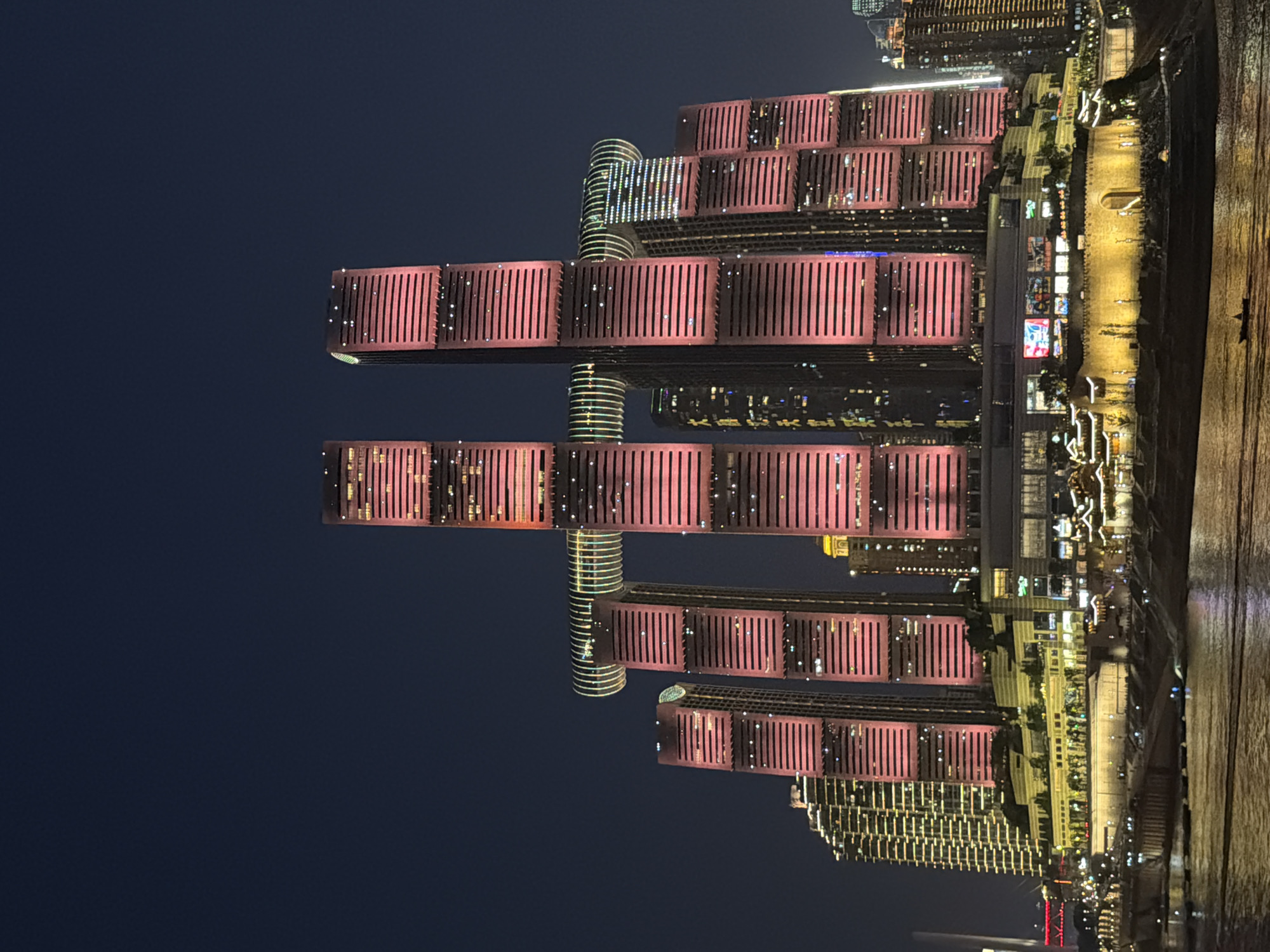 The image displays a nighttime cityscape dominated by the illuminated Raffles City Chongqing complex, situated in Yuzhong District but viewed from a vantage point across the Yangtze River, likely within the Nan'an District of Chongqing, China. Eight high-rise towers, arranged in a roughly rectangular formation, constitute the core of the complex. Most of these towers are illuminated with a striking red-pink light, emphasizing their horizontal architectural elements. A prominent multi-story horizontal skybridge, known as "The Crystal," connects four of the central towers near their upper sections, displaying segmented lighting that includes green, blue, and white hues.
The sky above is a uniform, dark indigo-blue, devoid of visible clouds or celestial objects. In the foreground, a wide body of water, presumed to be the Yangtze River, reflects the city lights. A small, dark boat with at least one occupant is visible on the water in the lower-right quadrant. Along the riverbank below the main complex, several illuminated structures are present. These include a large, golden-lit stone wall with a central arched entrance, flanked by traditional Chinese-style buildings featuring tiered, ornate roofs. To the left of the main complex, a cluster of modern buildings with various window lights is also visible. Further city lights and building outlines extend into the background on either side of the central structure.