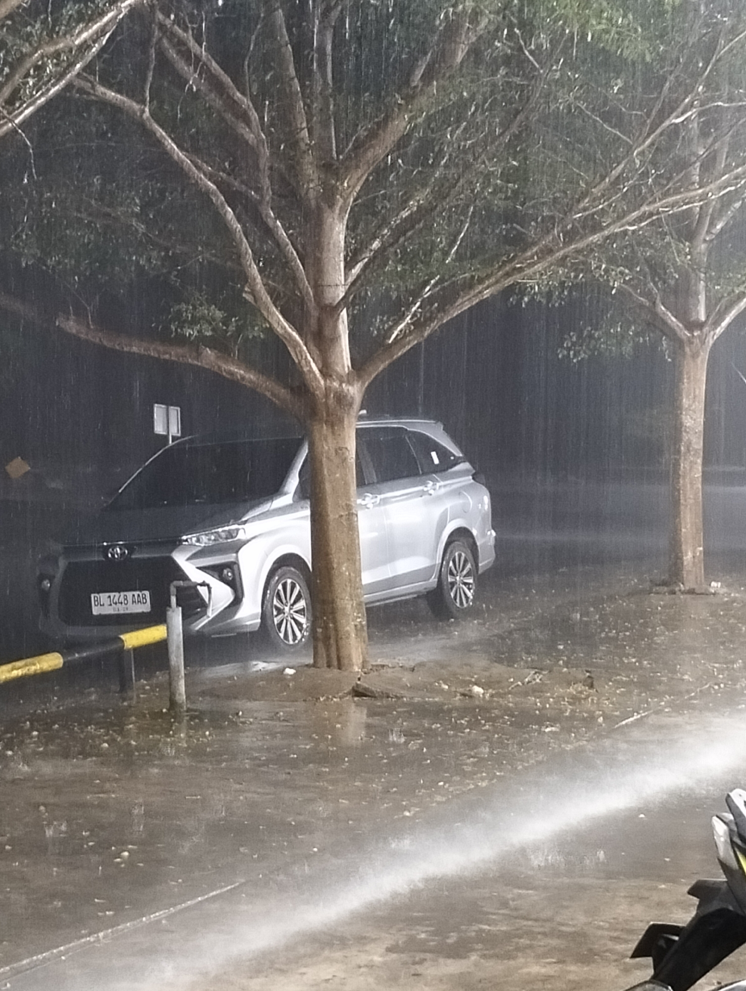 A silver Toyota Avanza is parked on a wet surface in Lhokseumawe, Indonesia, adjacent to a tree. It is raining heavily. The license plate reads "BL 1448 AAB". A yellow and black metal barrier is visible to the left of the vehicle. A second tree is seen in the background. A motorcycle is partially visible in the lower right corner of the frame.