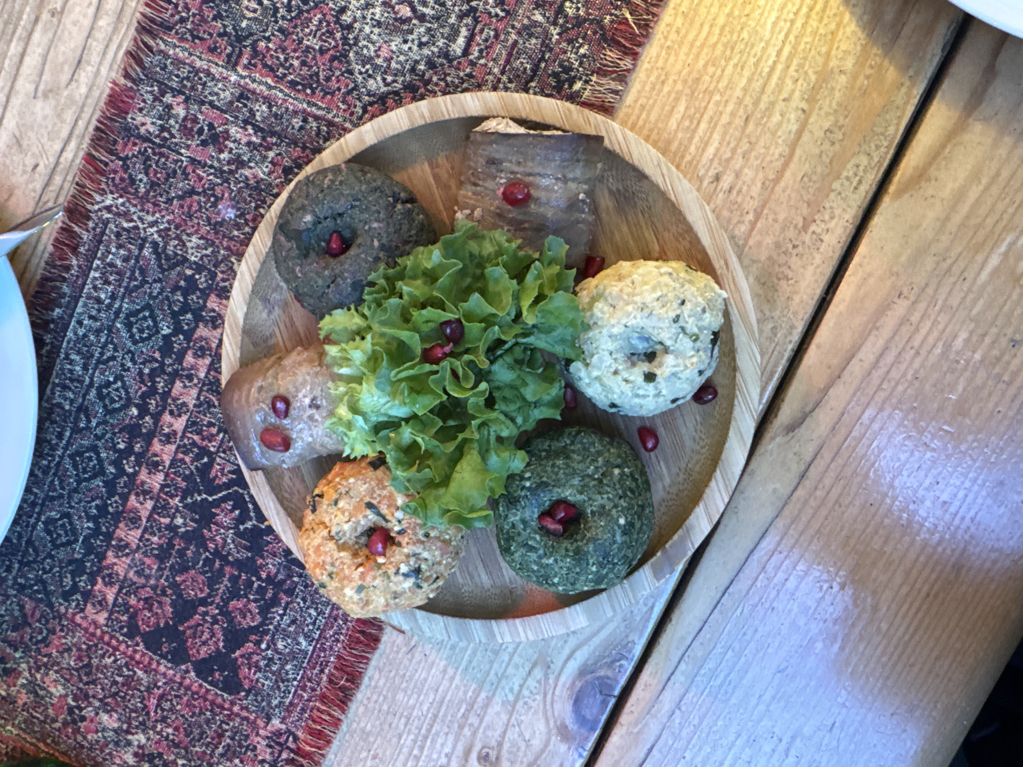 A bird's-eye-view, slightly-angled shot shows a wooden bowl containing several balls of Georgian food called pkhali on a wooden table in Tbilisi, Georgia. Pkhali are arranged around a mound of green lettuce with pomegranate seeds scattered throughout. The balls are various colors, including dark green, light brown, tan, white, and dark brown. Each ball has a small depression on top filled with pomegranate seeds. The wooden table is made of light-colored planks running vertically. A rug with a red and black geometric pattern is partially visible on the left side of the table, near a white plate.