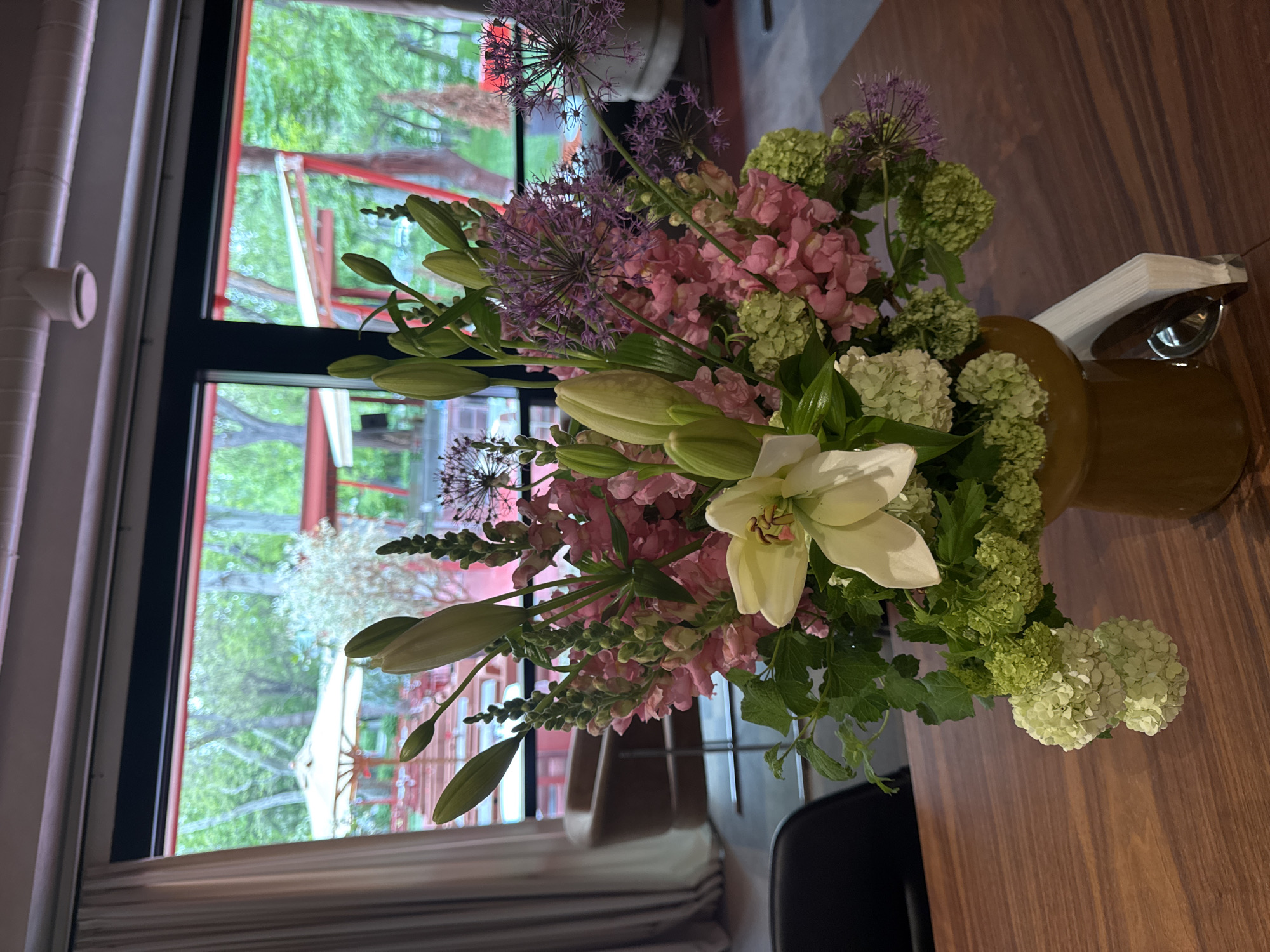 A vase filled with flowers sits on a wooden table in front of a window. The flowers are predominantly pink and purple, with white lilies and green hydrangeas interspersed. The vase is a dark golden brown color. A napkin holder and metal container with spoons or utensils are placed next to the vase. The window shows a view of trees and possibly a building with red supports outside. This image was likely taken indoors. It is not possible to determine the location within Yerevan, Armenia from the visual data provided.