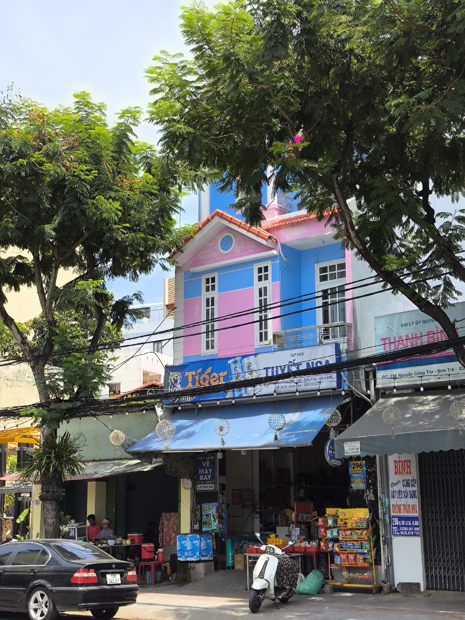 A street-level view of a vibrant commercial building in Da Nang, Vietnam, is captured on a bright, overcast day. The building's facade is painted in alternating horizontal stripes of bright pink and light blue, with a contrasting orange tiled roof. A white-framed window with multiple panes is visible on the second floor. Beneath this, a blue awning stretches across the storefront, featuring the "Tiger" logo with a tiger graphic, suggesting it may be a bar or restaurant. Above the awning, more signs are present, one in Vietnamese mentioning "TƯỢT NGA".
The street in front of the building is lined with power lines and decorative hanging lanterns. To the left, a black sedan is parked on the asphalt. To the right of the building, another storefront displays various goods, including snacks and drinks on a yellow display rack. A white scooter is parked in front of this shop, with a green bag placed behind it. Two individuals are visible inside the shop on the left, appearing to be attending to customers or managing the store. The environment suggests a typical bustling commercial street scene in a Vietnamese city, with a mix of residential and commercial activity.
