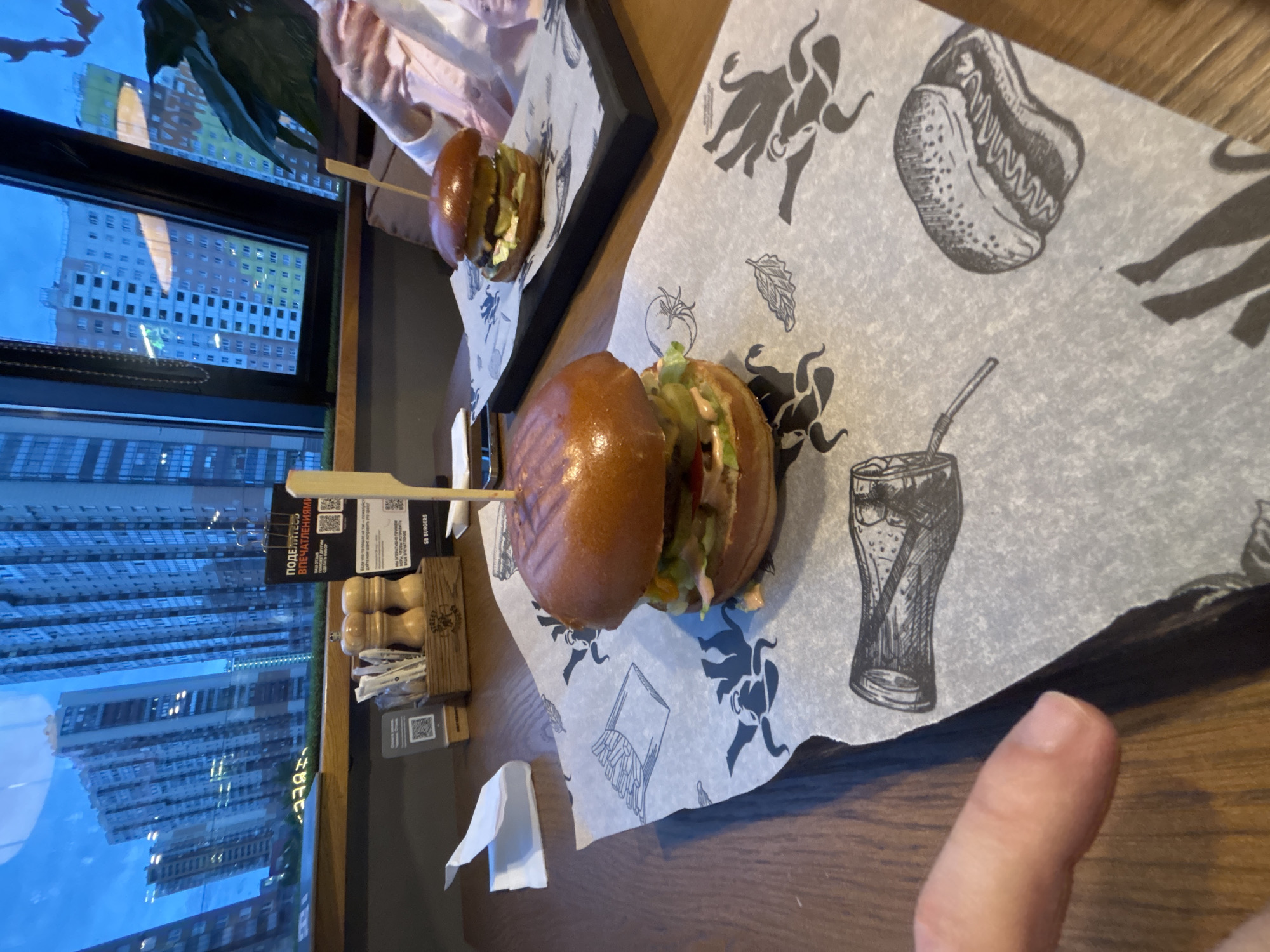 Here is a detailed description of the image:
**Scene Overview:**
The image depicts a restaurant setting with a view of an urban landscape through a window. A table is set with food, including burgers. The location appears to be Saint Petersburg, Russia, based on visible signs and architectural styles in the window view.
**Objects:**
* **Food:** Two burgers are prominently displayed on printed paper. One burger is larger and closer to the viewer, while the other is smaller and positioned further back.
* **Printed Paper:** The food is placed on sheets of paper with printed illustrations, likely serving as placemats or serving liners. The drawings include images of bulls, french fries, and drinks.
* **Table:** The food is set on a wooden table.
* **Condiments:** A small container holding salt and pepper shakers is visible near the window.
* **Napkin:** A white napkin is placed on the table.
* **Window:** A window shows a view of buildings, indicating an urban setting. The architecture and signs (in Russian) suggest the location is Saint Petersburg, Russia.
**Environment:**
* The image is taken indoors, likely in a restaurant or eatery.
* The natural light coming through the window suggests daytime.
* The setting appears modern and possibly trendy, based on the presentation of the food.
**People:**
* A finger is visible in the lower right corner of the image, suggesting that a person is holding a device used to capture the picture.
**Actions:**
* Someone is presumably dining at the restaurant, given the presence of the food and setting.
* The image appears to be taken by someone at the table.
**Interactions:**
* The burgers are presented on the table, ready to be consumed.
**Forensic Details:**
* The images is in good focus.
* The perspective is a slightly high angle.
* The lighting seems natural.