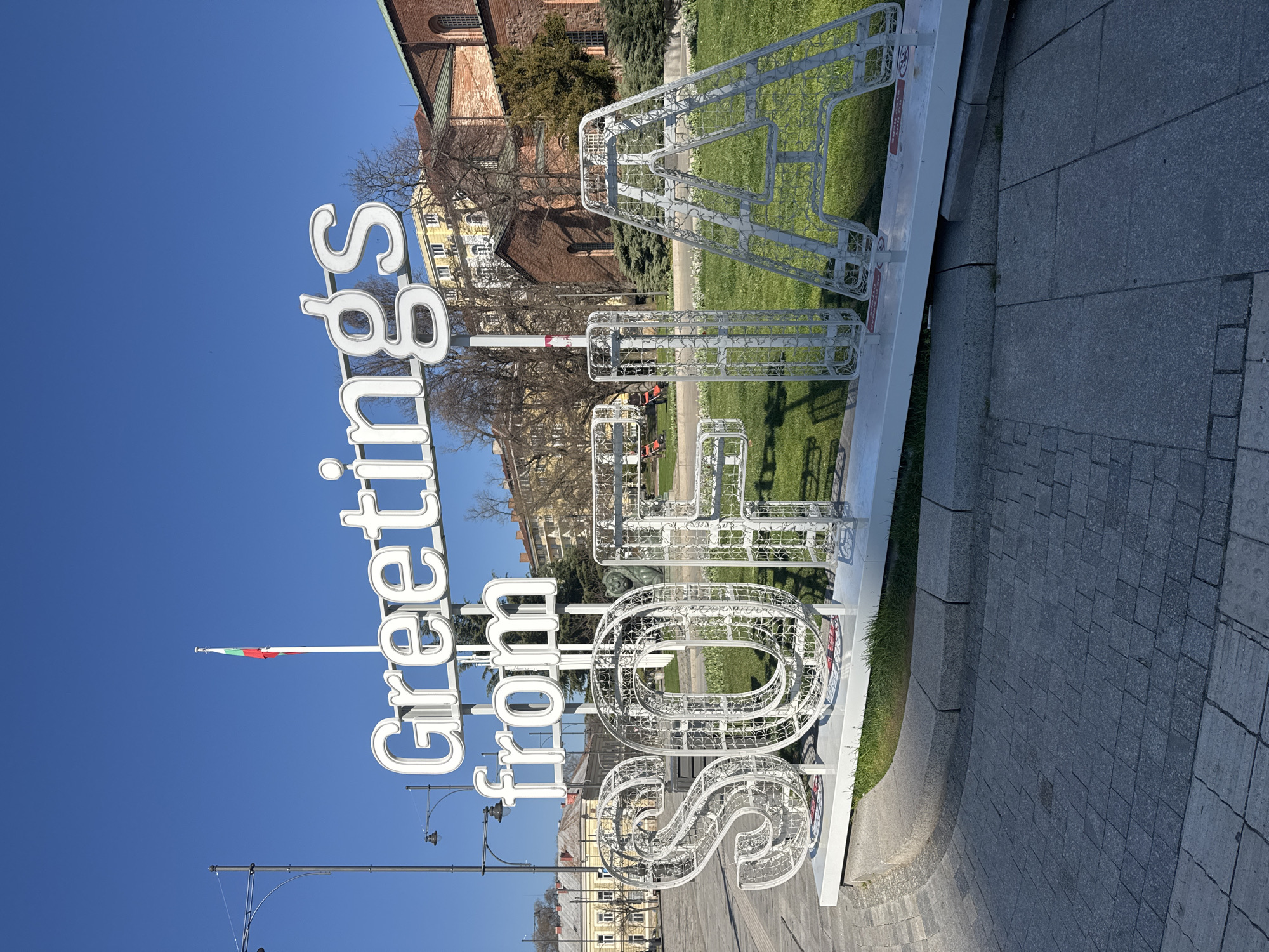The image depicts a large, white, illuminated sign that reads "Greetings from Sofia" in Sofia, Bulgaria. The sign is placed on a stone surface adjacent to a grassy area. A flag pole displaying the Bulgarian flag is visible. Buildings and trees are in the background. The sky is clear and blue.