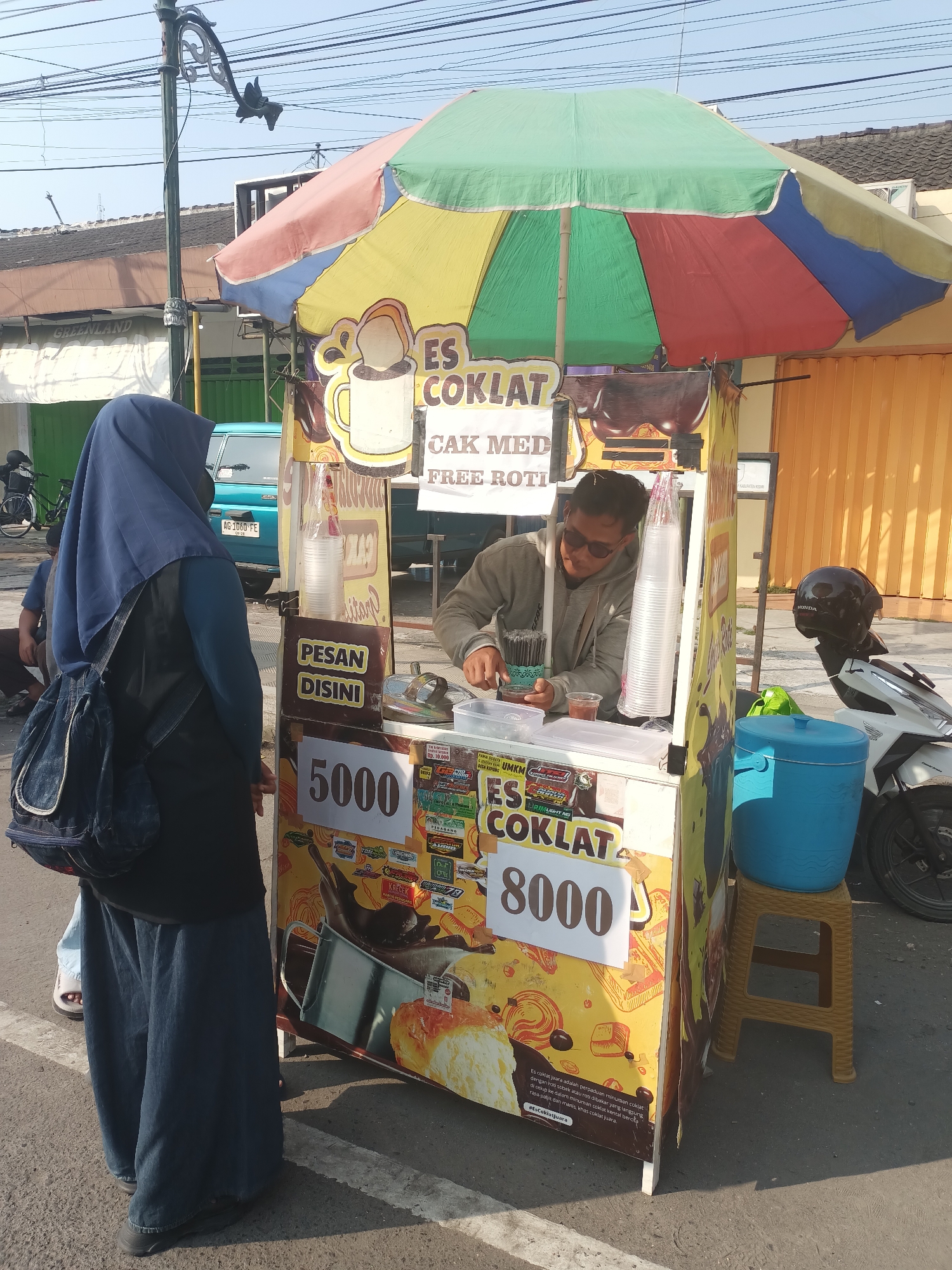 In the city of Pare, Indonesia, a vendor stands behind a colorful food cart selling "Es Coklat" (Chocolate Ice). The cart is adorned with images of chocolate and prices, with "5000" and "8000" prominently displayed. A customer, a woman wearing a blue hijab and denim clothing, is facing the cart. The vendor, a man with sunglasses, is preparing a drink. Several cups are stacked on the cart. A blue cooler sits on a yellow stool next to the cart, and a motorcycle is parked behind it. The scene is set on a street with a building and utility lines in the background.