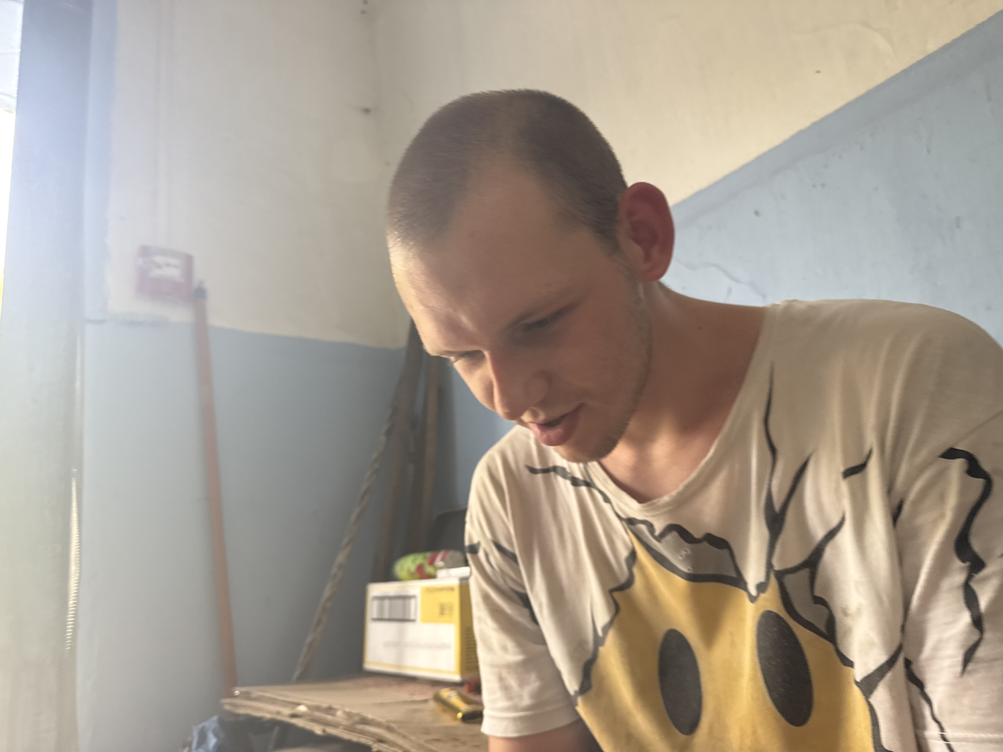 A young adult male with a shaved head and light stubble is looking down and to his right, his mouth slightly open. He is wearing a white t-shirt with a large yellow smiley face graphic on it. The background appears to be an interior space with light blue and white walls. To the left, a red fire alarm box is mounted on the wall. A long, thin wooden pole is visible next to the alarm. In the mid-ground, a cardboard box with writing on it is stacked. The lighting suggests it is daytime, with a strong light source coming from the left, creating a glare. The overall environment appears somewhat utilitarian and possibly industrial or institutional.