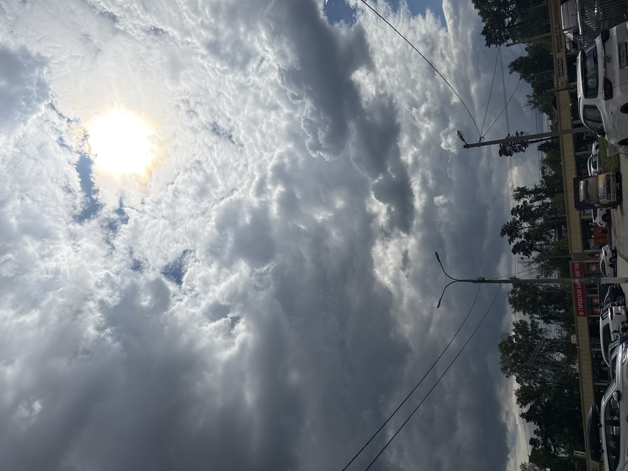 The image shows a view looking up at a cloudy sky with the sun partially obscured by the clouds. At the lower right edge of the frame, there are buildings, trees, and parked cars. The sign "ГОРОДСКАЯ БАЗА ЦВЕТОВ" (City Flower Base) is visible on a building, suggesting a flower shop is nearby. There are also streetlights and overhead power lines present. The location is likely in Mytishchi, Russia, given the user's context. The image captures an urban scene with a dramatic sky.