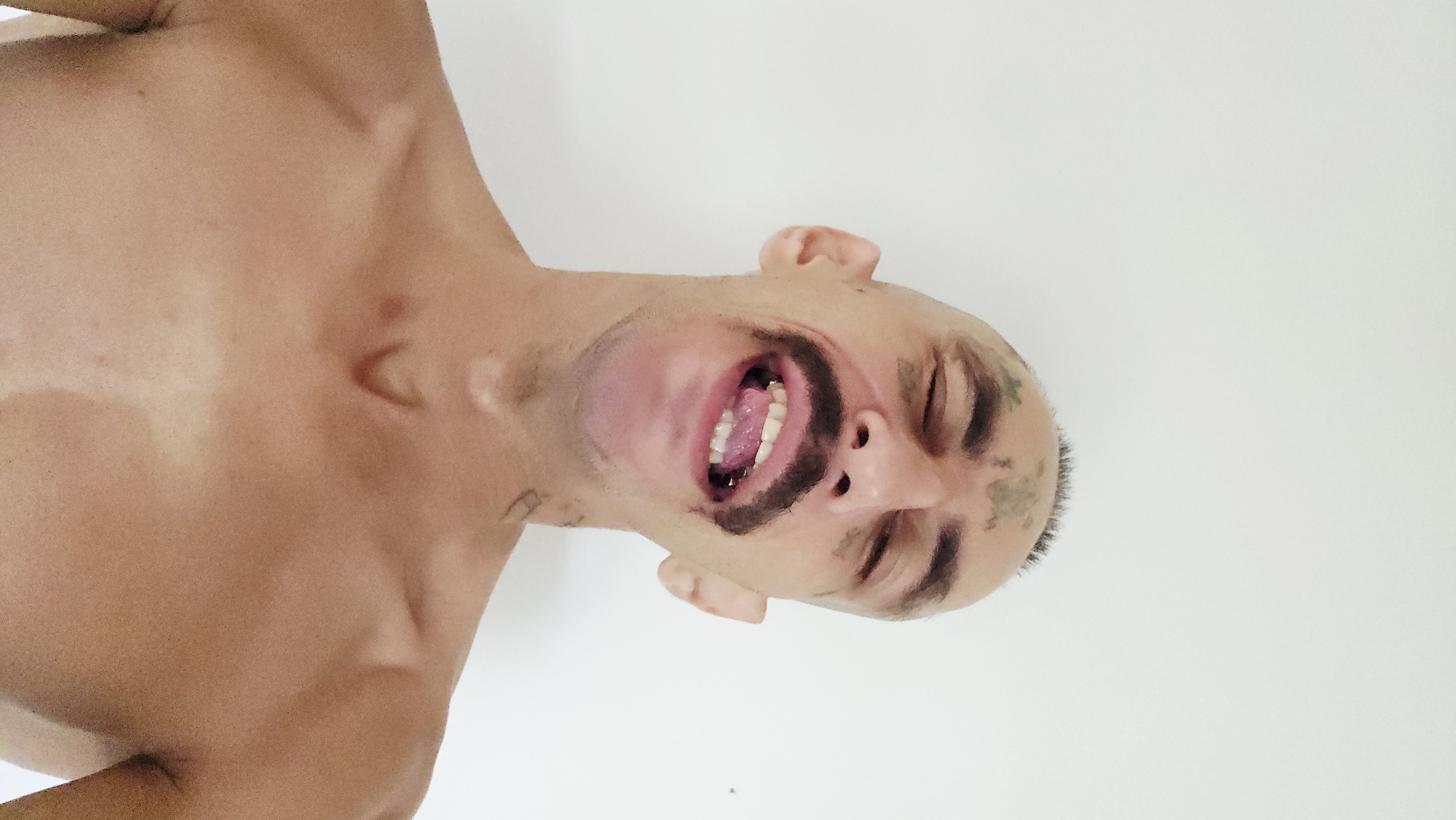 A medium-shot portrait depicts an adult person, appearing male, shirtless from the chest up against a plain white background. The image is oriented with the subject's head tilted upwards and to the right, as if looking towards the top of the frame.
The subject has short hair, possibly shaved, and striking dark, smudged markings resembling paint or makeup on their face. These markings delineate the chin, form a thick mustache-like shape above the upper lip, and extend across the temples and forehead. Faint, possibly tattooed, designs are visible on the temples/side of the head and a subtle marking on the neck.
The person's mouth is wide open, revealing white teeth and a pink tongue, while their eyes appear closed or tightly squinted. This creates an exaggerated, expressive look that could indicate a scream, intense laughter, or a dramatic pose. The bare chest and shoulders show a distinct tan line or area of lighter skin across the upper chest.
The scene is brightly and evenly lit, suggesting an indoor setting with a simple backdrop. The activity is a staged pose, possibly for performance or artistic photography. No other objects are present, and no text is visible in the image. The location context provided is Cam Lâm, Vietnam.