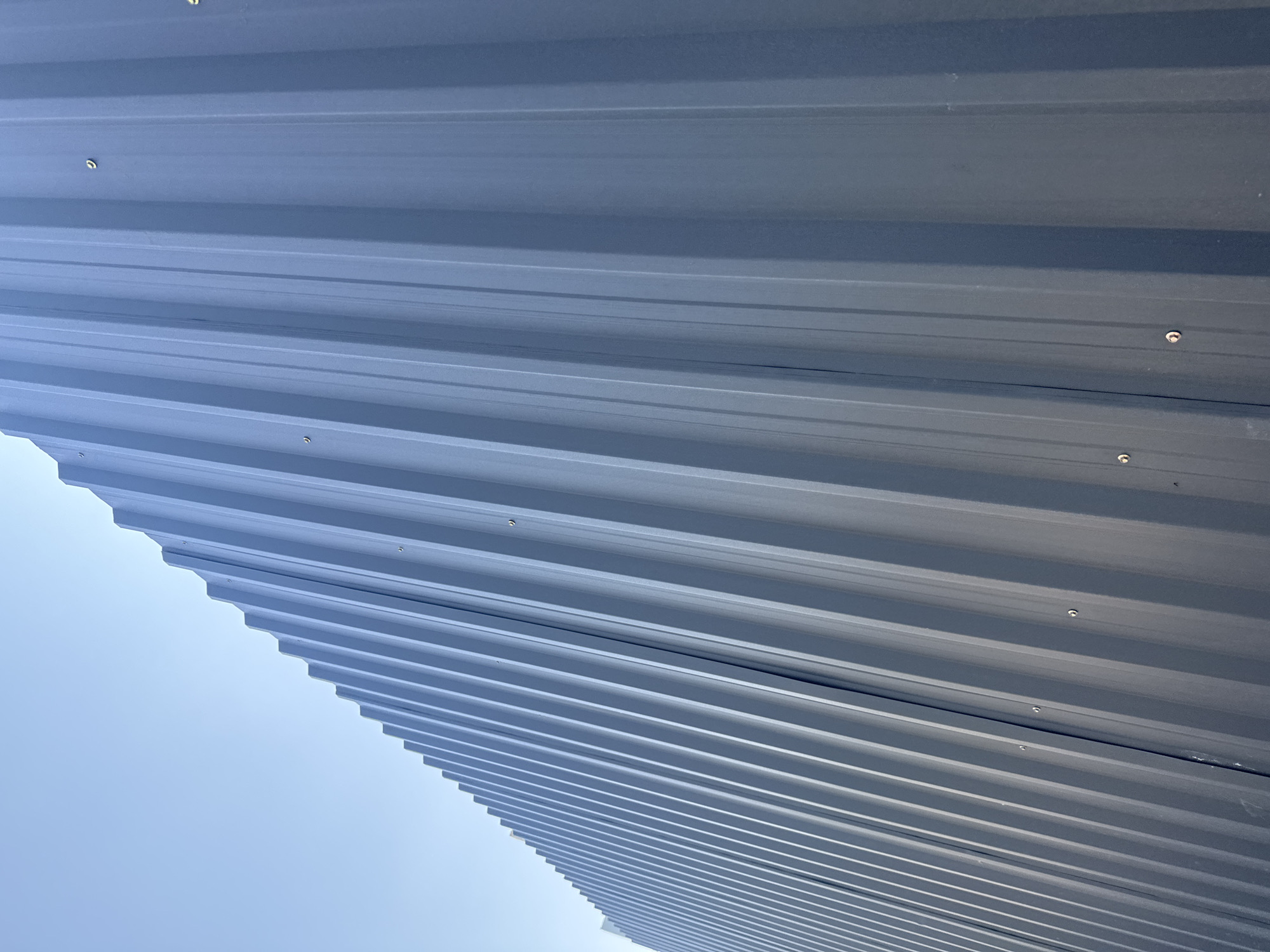 A low-angle, close-up shot of a dark grey corrugated metal wall with a clear blue sky in the background. The wall is made of vertical panels with a ribbed texture, creating a pattern of light and shadow. Several small screws are visible on the surface of the wall, securing the panels. The image appears to be taken outdoors, likely on a sunny day, given the bright sky and the strong highlights on the metal. The scene is devoid of any people or other distinguishing features, focusing solely on the architectural element.