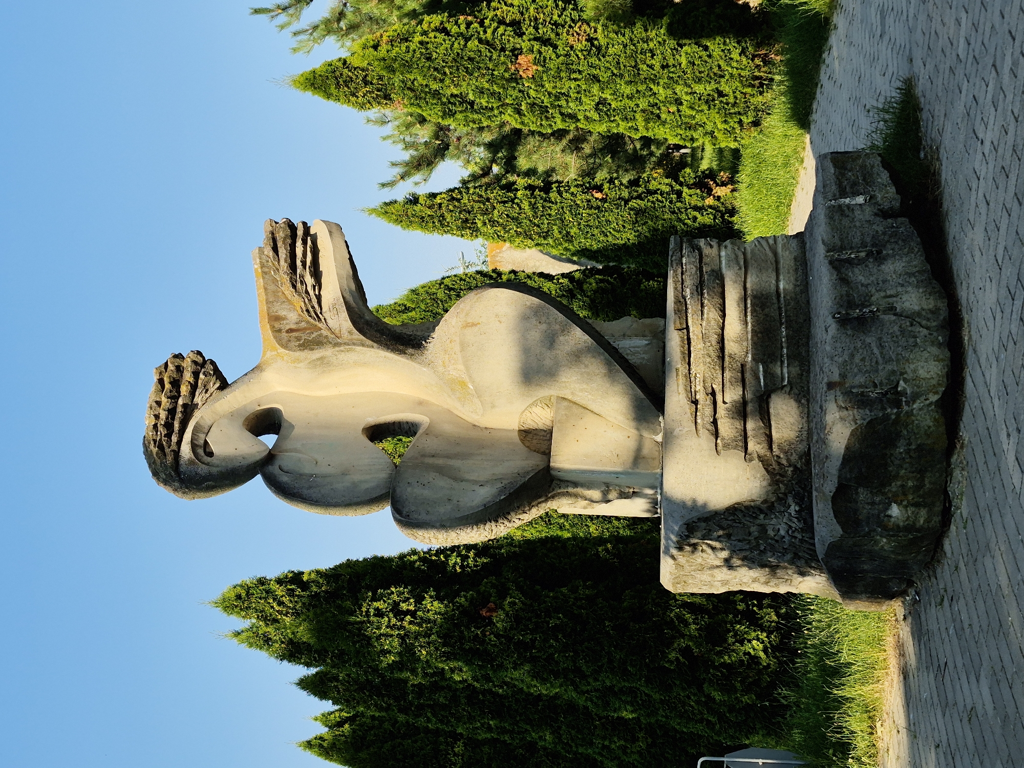 The image shows a large, abstract concrete sculpture of a stylized figure. The sculpture is light-colored and has a rough texture. It is positioned on a base made of layered stones, which in turn rests on a large, irregular boulder. The sculpture appears to be located outdoors in a park or public space. Behind the sculpture, several tall, conical evergreen trees are visible, some with patches of dried foliage. The ground in front of the sculpture is paved with light-colored bricks arranged in a pattern. A paved path curves to the right of the sculpture, bordered by green grass. The sky above is a clear, bright blue. The lighting suggests it is daytime, possibly late afternoon, with distinct shadows cast by the sculpture and trees. This sculpture is located in the "Sculpture Park" within the "Moldexpo" exhibition center in Chișinău, Moldova. There are no people or animals visible in the image.