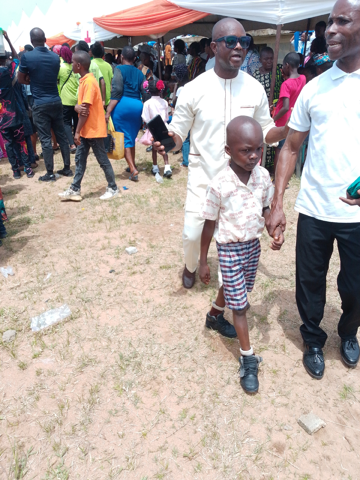 The image captures a scene in what appears to be Abak, Nigeria, judging by the people and environmental elements.
The foreground is dominated by a young boy in a shirt with school-related text, and plaid shorts. He is holding hands with two adult men; one wears sunglasses and an off-white tunic-style shirt with matching pants, and the other wears a white polo shirt and black pants.
In the background, there is a large group of people gathered under a makeshift awning constructed with orange and white fabric. People are wearing a variety of clothing, including t-shirts, skirts, and traditional attire.
The ground is dry and partially covered with sparse vegetation.