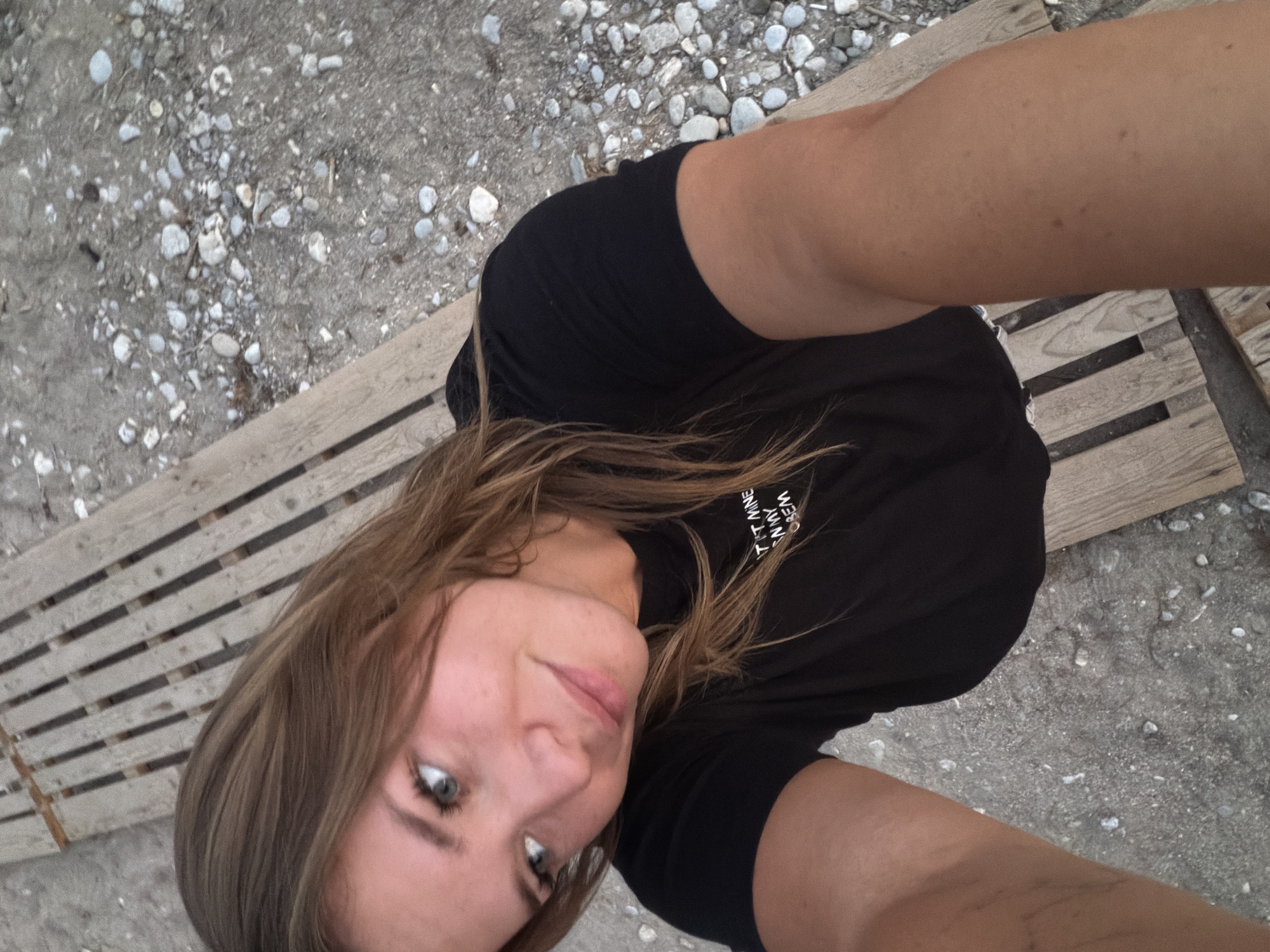 A young woman with light brown hair is taking a selfie outdoors. She is wearing a black t-shirt with white text on it. She is sitting on a wooden bench on a gravel beach. The image appears to have been taken during the day, as there is natural light. The location is the city of Prijevor, Montenegro.