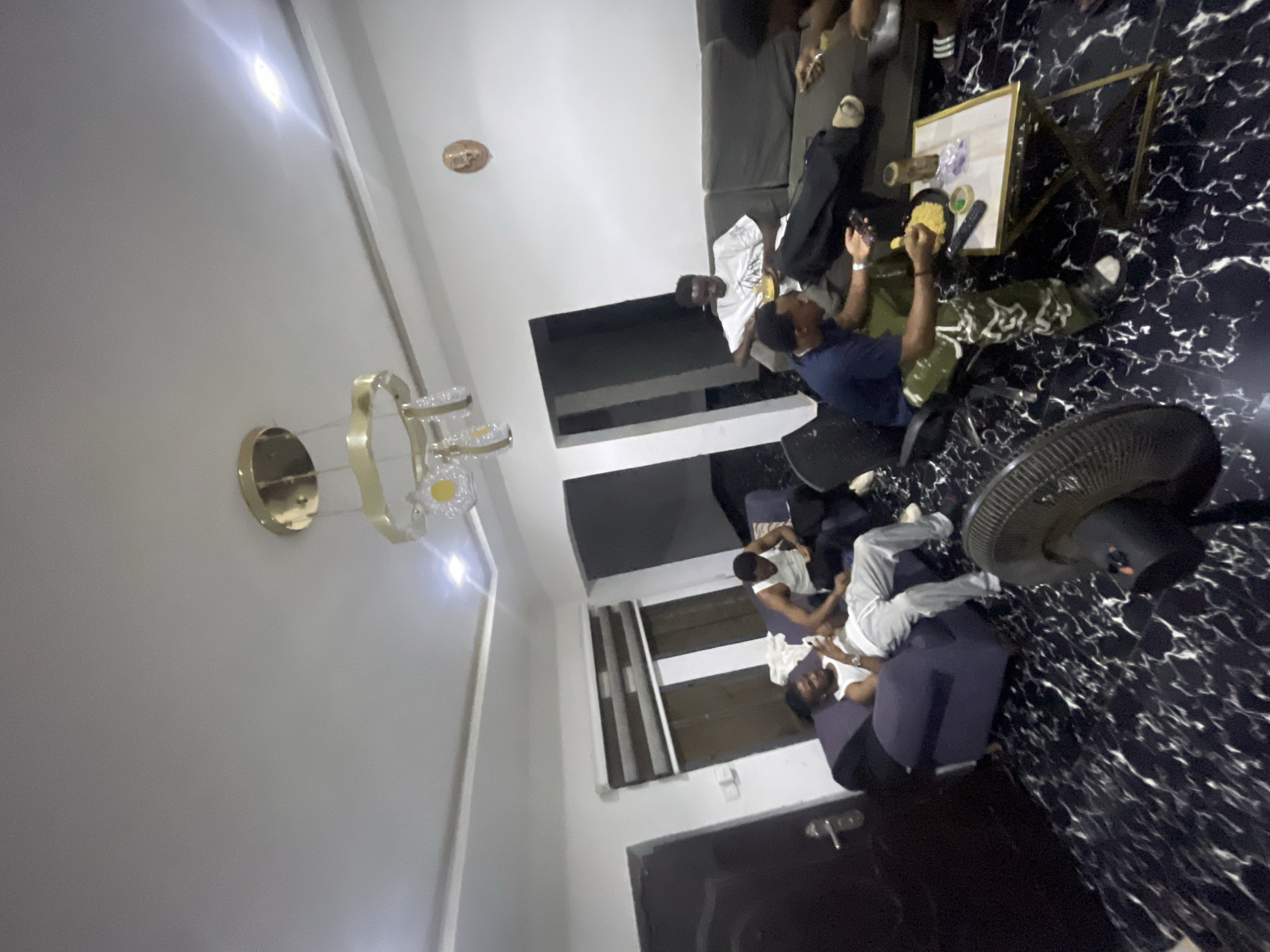 The image depicts an interior space, likely a living room, in Abuja, Nigeria. There are multiple individuals present, engaged in various activities. Some are seated on a dark grey sectional sofa, while others are seated on blue armchairs. A person is standing, appearing to interact with someone seated. The floor has a glossy, marbled black and white pattern. A ceiling fan is visible in the foreground. A low table with objects on it is present. A chandelier-style light fixture is mounted on the wall.