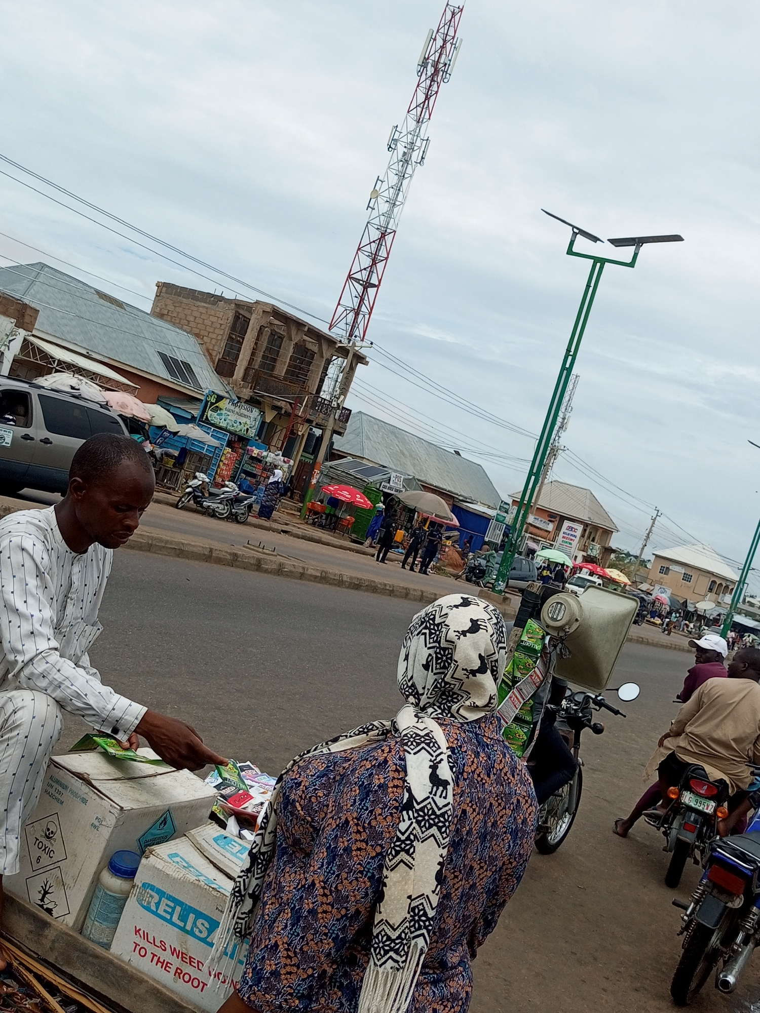 A man in a striped shirt gestures towards a box with his left hand, offering items to a woman wearing a headscarf and a patterned dress. The box, labeled "RELISA KILLS WEED TO THE ROOT," contains various small packets. To the left of the box is another box marked with a "TOXIC" symbol. The scene takes place on a paved street next to a sidewalk lined with market stalls and buildings. In the background, a tall red and white telecommunications tower rises into the sky, accompanied by a green solar-powered street light. Several motorcycles are parked along the street, and other people are visible in the distance, some on motorcycles and others near the market stalls. The sky is overcast.