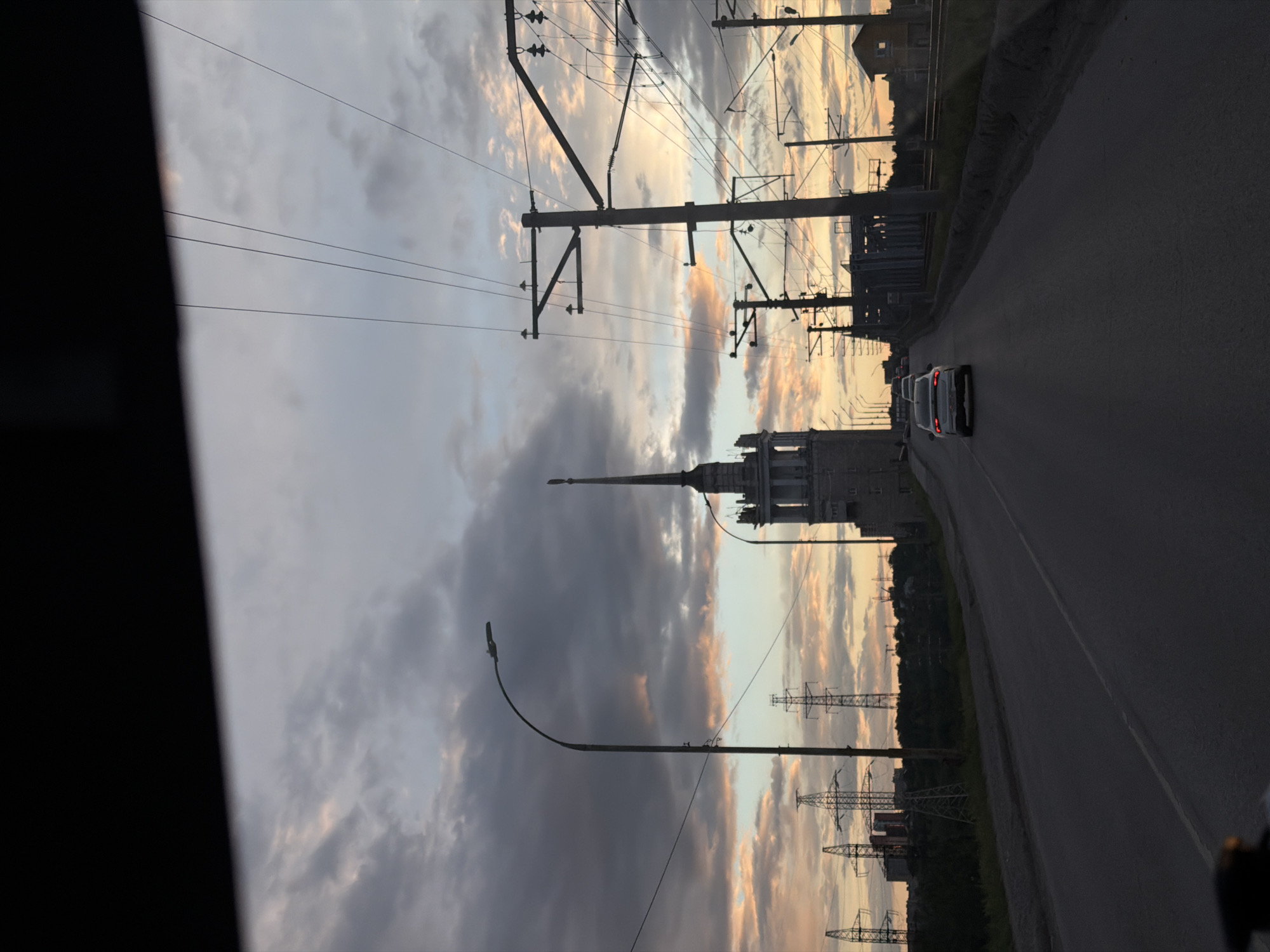 The image captures a twilight scene in Perm, Russia, notable for its urban and industrial elements. A road stretches into the distance, occupied by a line of vehicles. Overhead, power lines and supporting structures are prominent, silhouetted against a sky with a mix of dark clouds and patches of light, suggesting either sunrise or sunset. A tall, pointed building stands out amidst the industrial landscape, possibly a notable landmark of Perm. Streetlights line the road, indicating urban infrastructure. The presence of power lines and industrial structures suggests proximity to an industrial zone within the city. The view is likely taken from a moving vehicle.