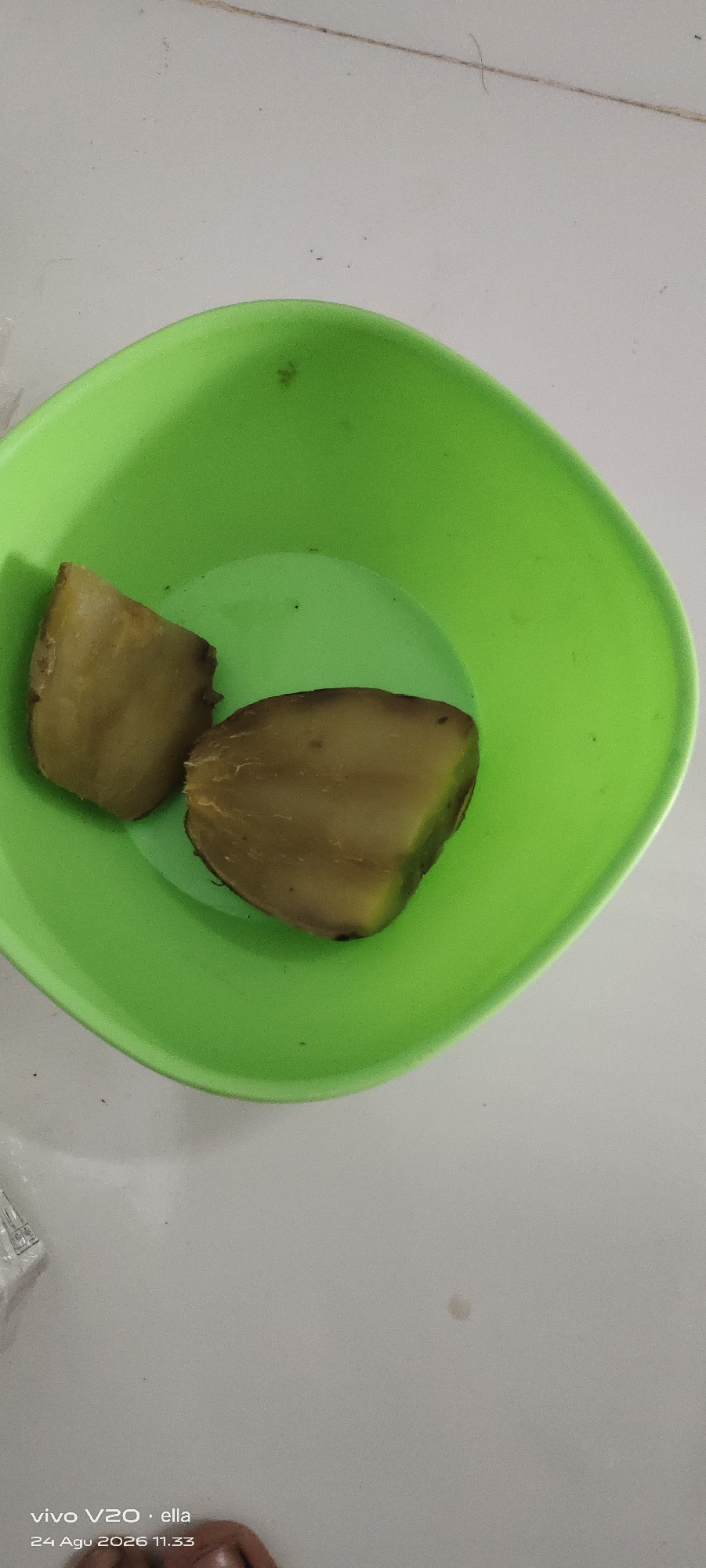 Two pieces of cooked potato are in a bright green plastic bowl on a white surface. The potatoes are a light brown color and appear to be soft. The bowl is rounded and has a wide rim. The surface the bowl is on is textured and appears to be a tabletop or counter. The timestamp at the bottom of the image indicates it was taken on August 24, 2026, at 11:33 AM in Simo, Indonesia. There are no people visible in the image.