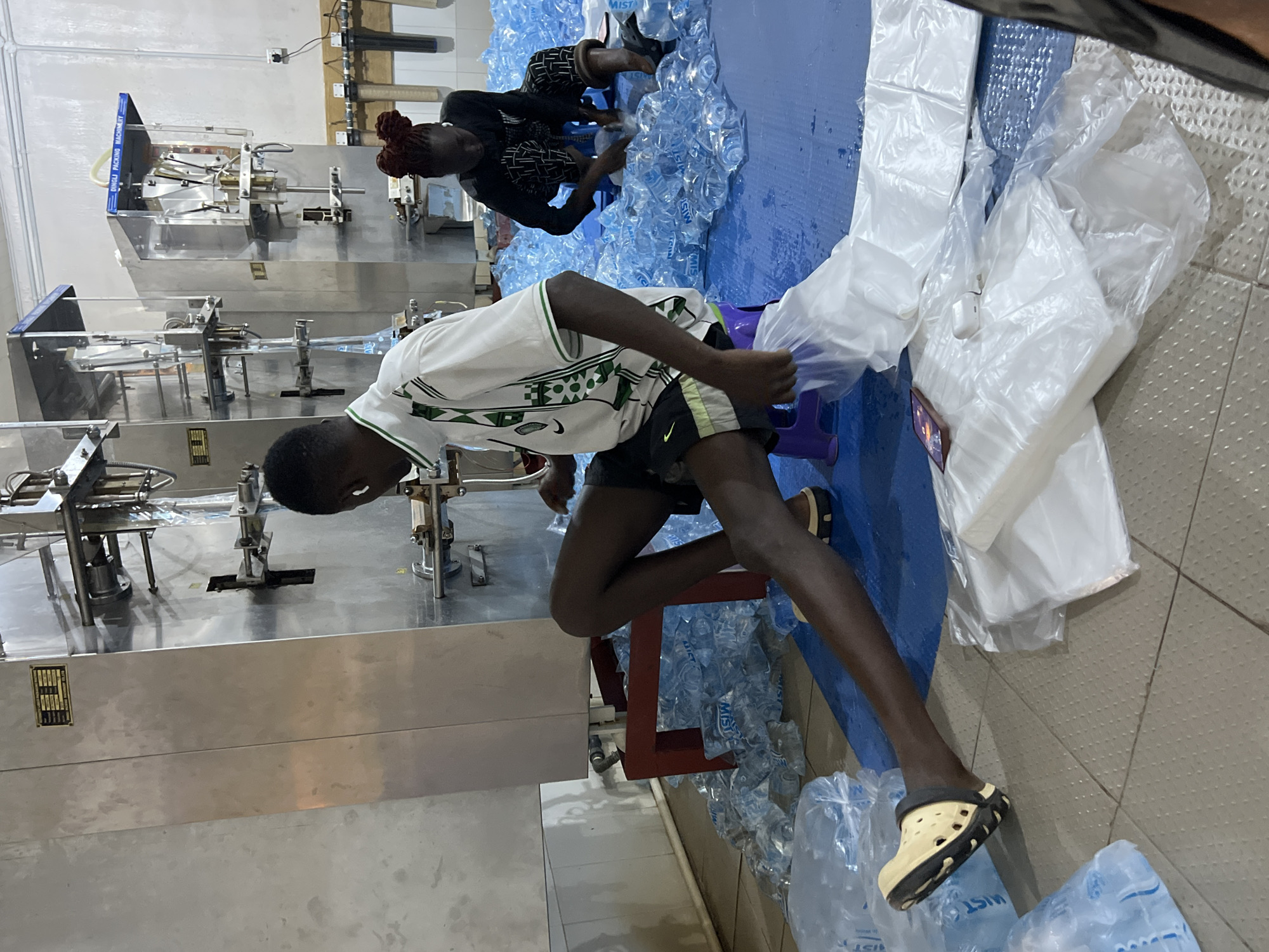 The image captures an indoor scene at what appears to be a water bottling plant in Jos, Nigeria. Two individuals are visible, seemingly involved in the bagging or packaging of bottled water. The bottling process appears to be automated, with a large silver machine on the left dispensing water bottles. One individual, wearing a white shirt with green and white details and black shorts, is placing bottles into a large plastic bag. The other individual, with red hair, is helping pack the water bottles. Numerous filled water bottles are scattered on the floor around the two individuals. The floor is tiled.
