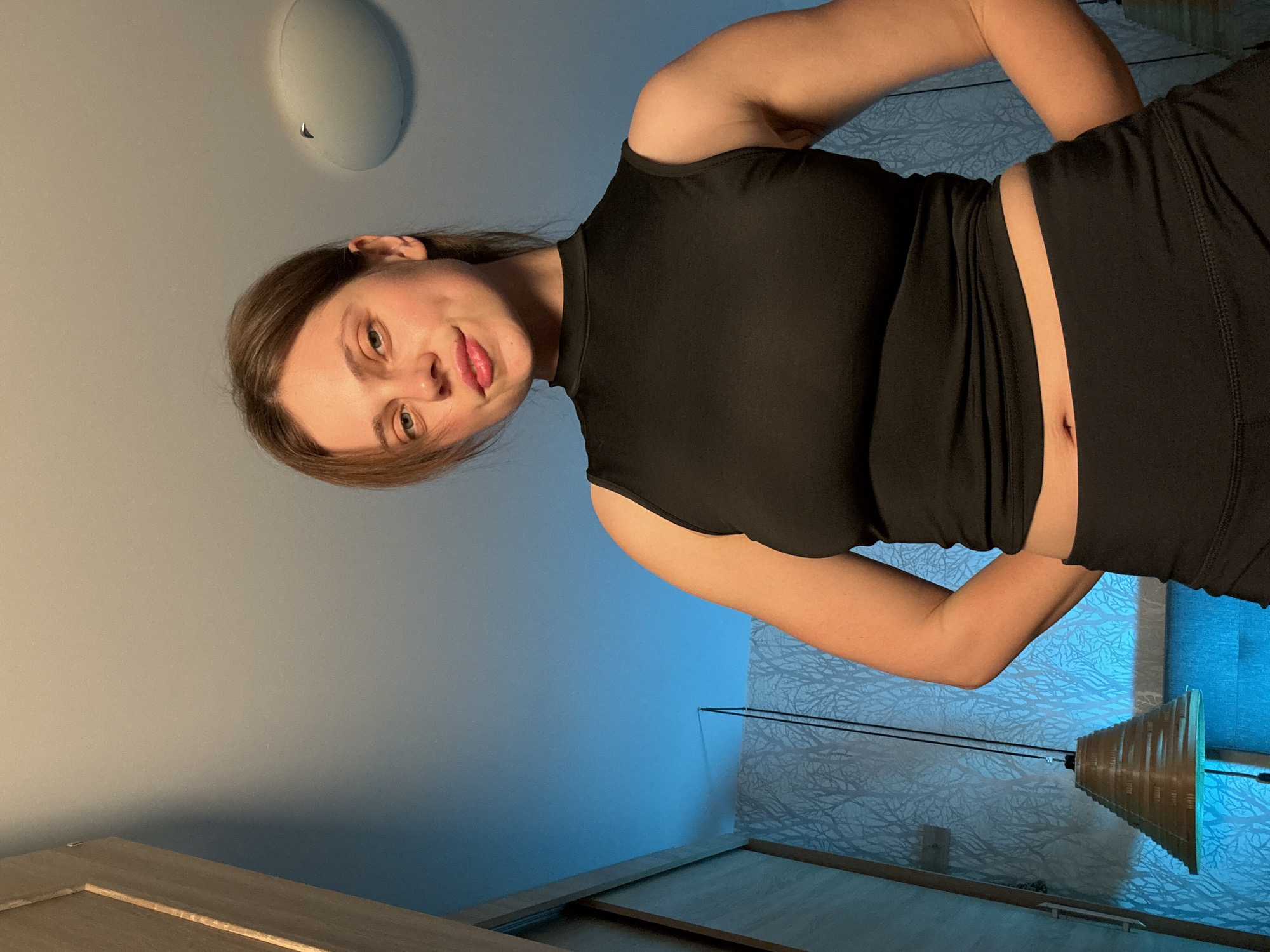 A young woman with light brown hair and fair skin is looking directly at the camera, her head tilted slightly. She is wearing a black sleeveless mock-neck top that exposes her midriff, and black bottoms. Her hands are clasped behind her back. The background is a wall with a light blue hue, adorned with a subtle tree branch pattern wallpaper. To the left of the frame, a wooden closet door is visible. A unique, conical lamp with horizontal slats hangs from the ceiling, casting a warm glow. The scene appears to be indoors, possibly in a residential setting. The photograph was taken in Vrchlabí, Czechia.