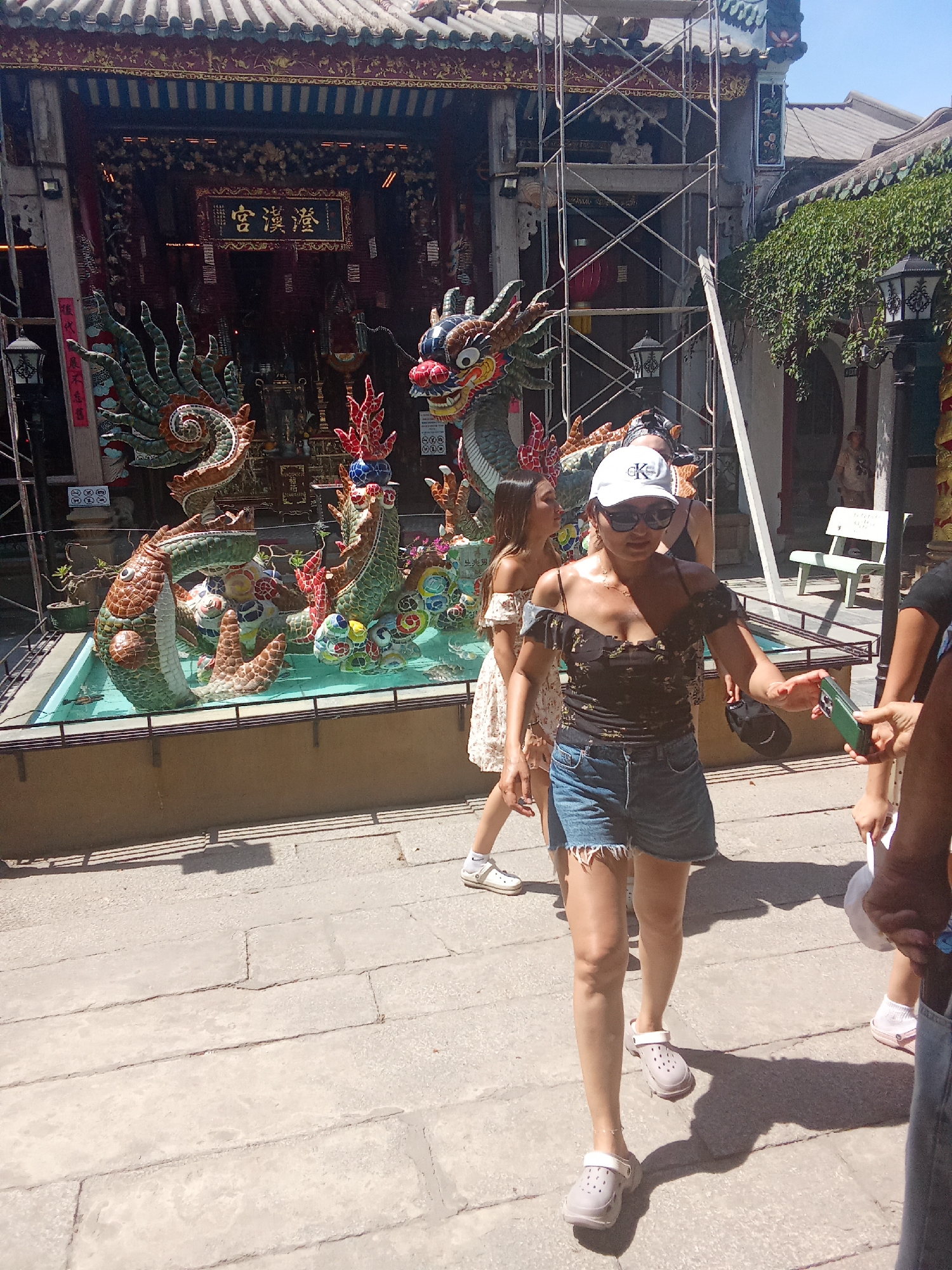 In Hội An Ward, Vietnam, a group of women are walking near a temple adorned with intricate dragon sculptures. In the foreground, a woman in a black floral off-the-shoulder top and denim shorts walks towards the camera, holding a green smartphone in her outstretched right hand. She is wearing a white baseball cap and sunglasses, and light-colored clogs. To her left, another woman in a white floral dress walks away from the camera. In the background, a temple structure with traditional Vietnamese architecture is visible, featuring a prominent sign with Chinese characters. Scaffolding is present on the right side of the temple, suggesting ongoing construction or maintenance. Dragon statues are displayed in a shallow pool in front of the temple. The ground is paved with stone slabs, and shadows from the surrounding structures create a dappled effect.