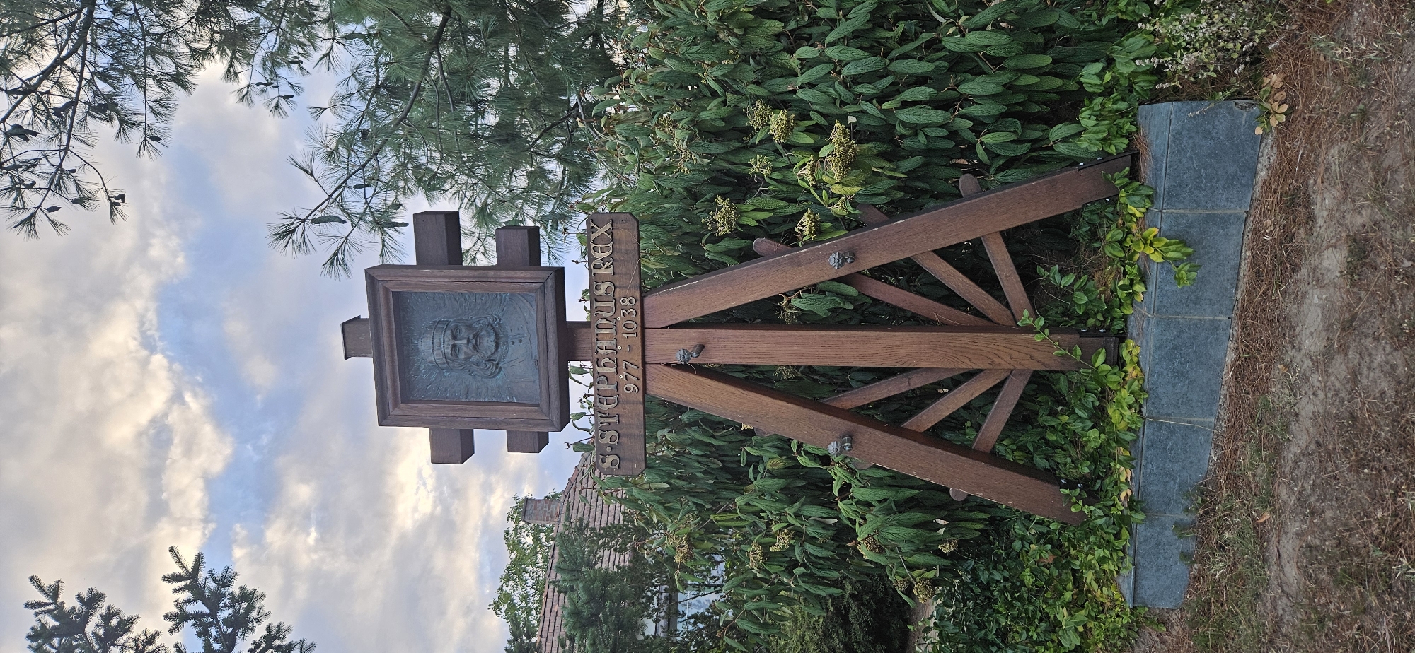 A vertical, eye-level shot depicts an outdoor memorial dedicated to Saint Stephen of Hungary in Hévíz, Hungary. The memorial consists of a wooden cross-shaped structure supporting a bronze bas-relief portrait of a man with a beard and crown, identified by the inscription below as "S. STEPHANUS REX 977-1038". The portrait is set within a dark wooden frame. The wooden structure is supported by a tripod-like base made of thicker wooden beams. The memorial is situated in front of a dense green shrubbery, with pine trees visible in the background and upper portions of the image. A small building with a tiled roof and a brick chimney can be seen to the left, peeking through the foliage. The ground in front of the memorial is covered with dry pine needles and dirt. The sky above is partly cloudy with patches of blue. The lighting suggests it is either early morning or late afternoon.