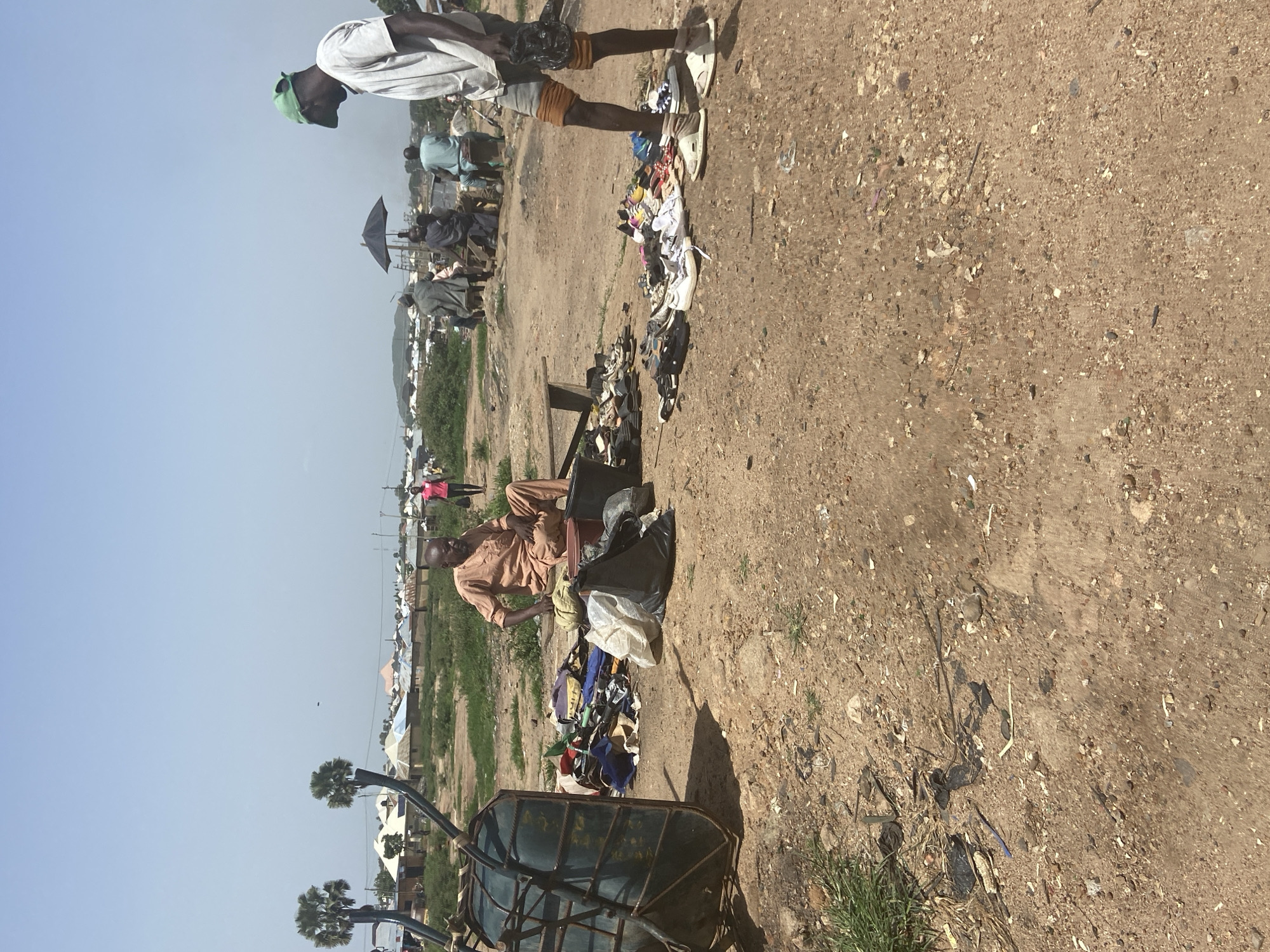 The image depicts an outdoor scene in Bukuru, Nigeria, with a clear blue sky, suggesting it is daytime. The ground is dry and dusty, with scattered debris, characteristic of an arid or semi-arid environment.
In the foreground, a man in a light brown shirt is seated on a chair or bench, with several black and white bags beside him. In front of him, a pile of clothing and various items is displayed on the ground. To the left, a metal trolley with a green-covered cage is partially visible.
Further back, another man, wearing a white t-shirt and orange shorts, stands with a green cap. He is holding a dark bag. Behind him, a group of people are seen, some gathered around, and an umbrella is open, providing shade.
In the background, there are indications of a settlement with various buildings, some with colorful roofs, and a stretch of green grass. The overall scene suggests a marketplace or a gathering area where goods, possibly second-hand clothing, are being sold or exchanged. The presence of a trolley and scattered items implies a commercial or informal trading activity. The setting appears to be a relatively open area within the community.