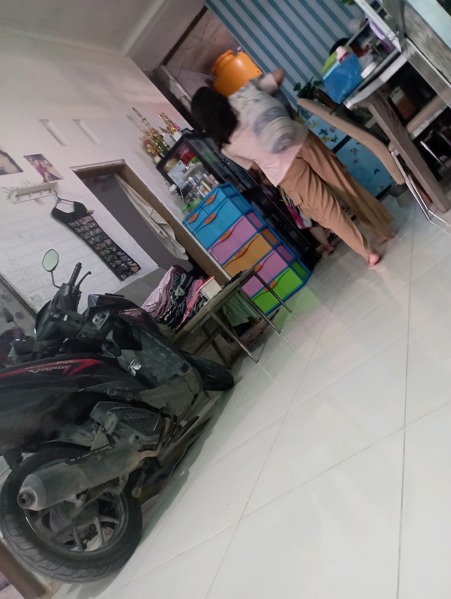 The image depicts an interior scene with a motorcycle in the foreground, parked on a tiled floor. A person, seen from the back and side, is standing and interacting with a colorful cube storage unit. The storage unit is composed of individual colored cubes, with visible shades of blue, orange, pink, and green. The walls are white, with some decorative elements like framed pictures and a hanging organizer for sunglasses. In the background, a shelf unit holds various items, including what appears to be trophies and bottles. The overall impression is of a cluttered domestic or storage space. The image does not provide specific visual cues to confirm the location within Ketapang Lor, Indonesia.