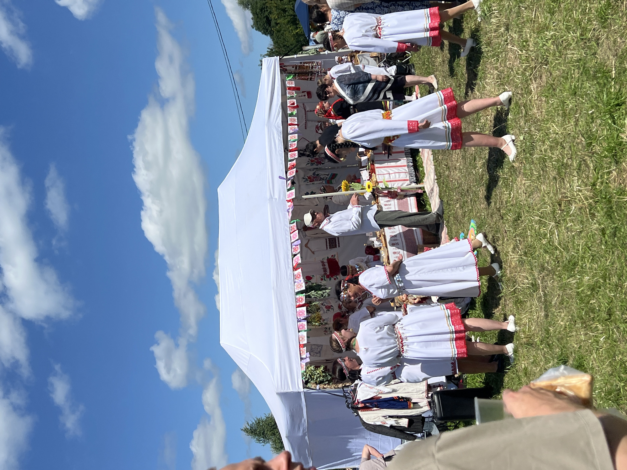 The image depicts an outdoor gathering under a clear blue sky with scattered white clouds, likely at a festival or market in Kalinino, Russia. A large white tent serves as a central structure, decorated with colorful, folkloric banners. Inside and around the tent, people are engaged in various activities.
Several individuals, predominantly women, are dressed in traditional white outfits adorned with red and patterned embroidery. Some are standing and conversing, while others appear to be interacting with a stall. One woman in a white dress with red trim is holding her hands up in a gesture of explanation or demonstration. Another group of women in similar attire are gathered, looking towards the center of the scene.
In front of the tent, a table is set up with various items, suggesting a sale or display of goods. These items are not clearly identifiable but appear to be small and varied. To the left of the tent, clothing items are displayed on a rack, including jackets and shirts with intricate embroidery.
A man in a white shirt and light-colored pants, wearing a cap, stands near the table, possibly attending to customers or presenting items. Another man, partially visible in the foreground and out of focus, is holding a clear plastic cup and a bag containing what appears to be food.
The ground is covered in green grass, indicating a natural or park-like setting. The overall atmosphere seems lively and festive, with a strong emphasis on cultural tradition, as evidenced by the attire and decorations.