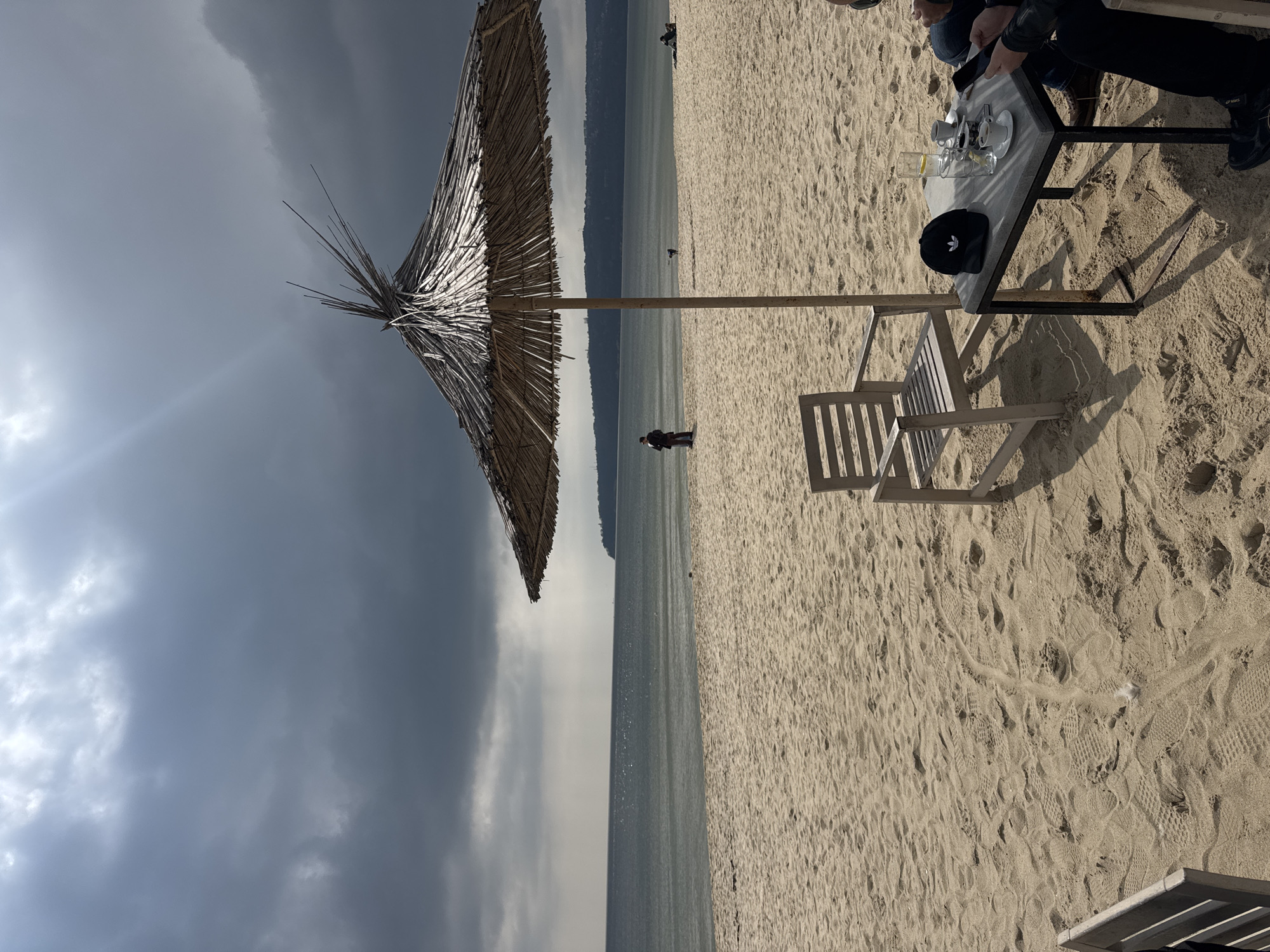 The image depicts a beach scene near Varna, Bulgaria, under a stormy sky. In the foreground, a thatched beach umbrella casts a shadow on the sand, next to a white wooden beach chair and a metal table with a black Adidas cap and coffee cups. Two people are partially visible at the table. In the background, a figure is walking alone on the beach near the shoreline, where a narrow strip of land meets the sea, fading into distant hills. The overall mood is subdued due to the overcast sky.