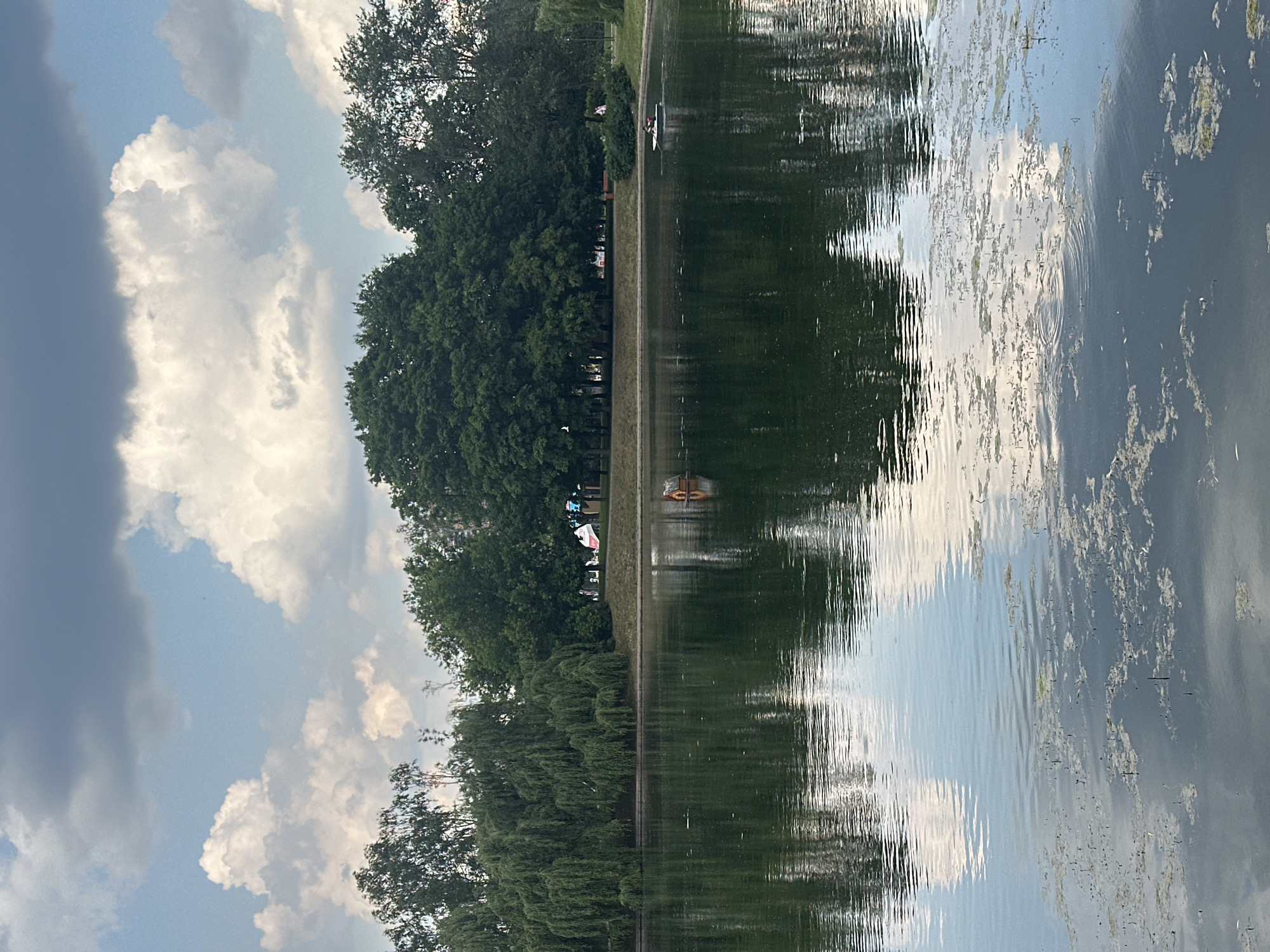 The image shows a serene lake in Khimki, Russia, with lush green trees lining its bank. The sky is partly cloudy, with fluffy white clouds contrasting against a blue sky. The water's surface reflects the surrounding trees and clouds, creating a beautiful, almost ethereal scene.
On the lake, a small wooden duck house floats near the left bank. Further out in the water, a person is seen in a small boat, enjoying a peaceful ride. In the distance, some trees are decorated with colorful flags, possibly indicating a local festival or event. The overall atmosphere is tranquil and inviting, a perfect spot for relaxation and recreation in the city.