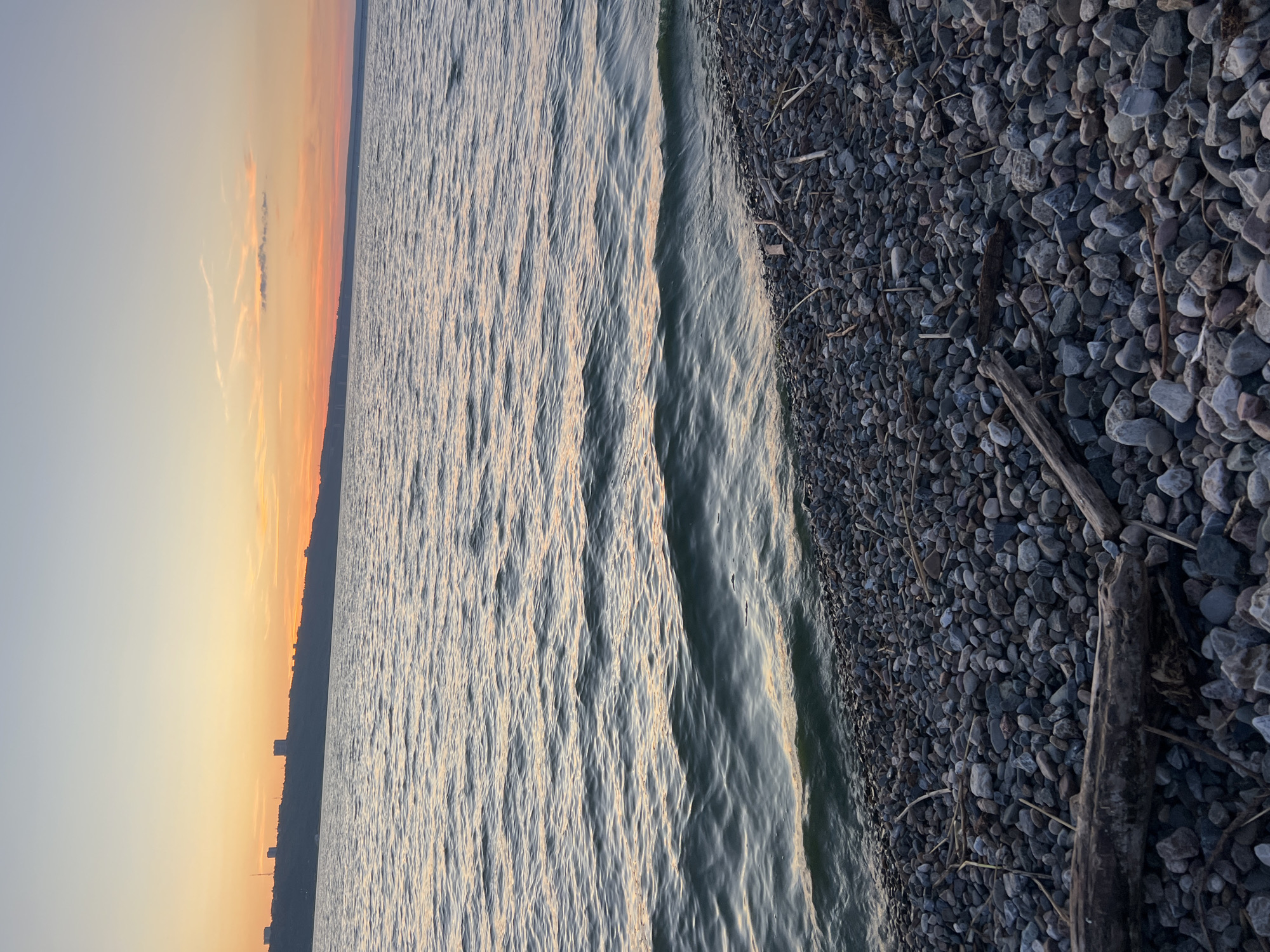 The image depicts a rocky shoreline of a body of water, likely a river or lake, with a city skyline visible on the distant horizon under a twilight sky. The foreground is dominated by a dense scattering of grey, smooth stones and driftwood. The water exhibits gentle ripples, reflecting the warm hues of the sunset, which casts an orange and pink glow across the sky. The city skyline appears as a dark silhouette against the lighter sky, punctuated by several tall, block-like structures, possibly apartment buildings or administrative centers. The scene suggests a tranquil evening at the edge of an urban area. Given Ulyanovsk's location on the Volga River, it is plausible this scene is captured along its embankment, perhaps near the Presidential Bridge or the residential areas overlooking the river.