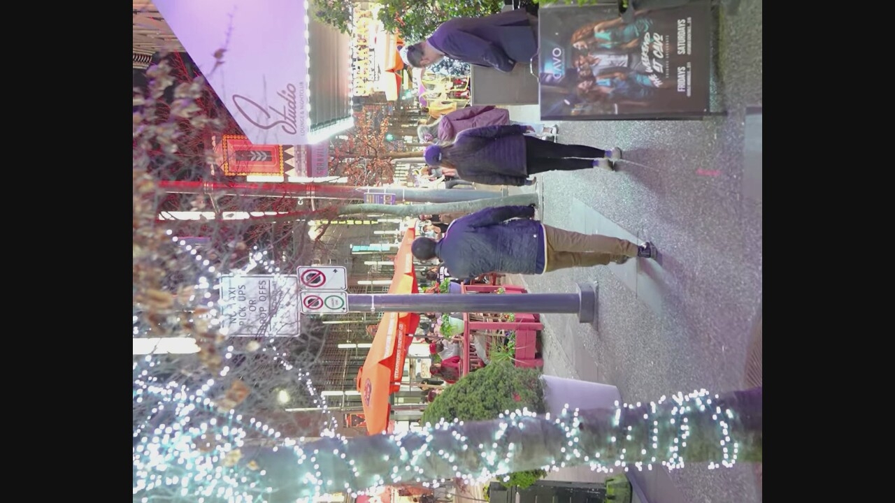 The image depicts a nighttime street scene in Vancouver, Canada, likely on Granville Street given the visible signage. The street is illuminated by decorative string lights on trees and neon signs from businesses. In the foreground, several people are walking on a sidewalk. One man in a dark jacket and brown pants is walking away from the camera. To his right, a woman wearing a blue puffer jacket and a purple beanie is walking in the same direction. Further down the sidewalk, another person in a dark jacket is partially visible.
On the left side of the image, an outdoor seating area for a restaurant or cafe is set up with orange umbrellas and red tables. Signs are visible on a pole, one indicating "NO TAXI PICK UPS OR DROP OFFS". Another sign for "Studio Lounge & Nightclub" is visible on the right side of the street, with "Granville" text also apparent. A poster on an easel on the right advertises "THE WEEKEND AT CAVO" with "FRIDAYS SATURDAYS" listed. The ground is paved with asphalt and concrete.