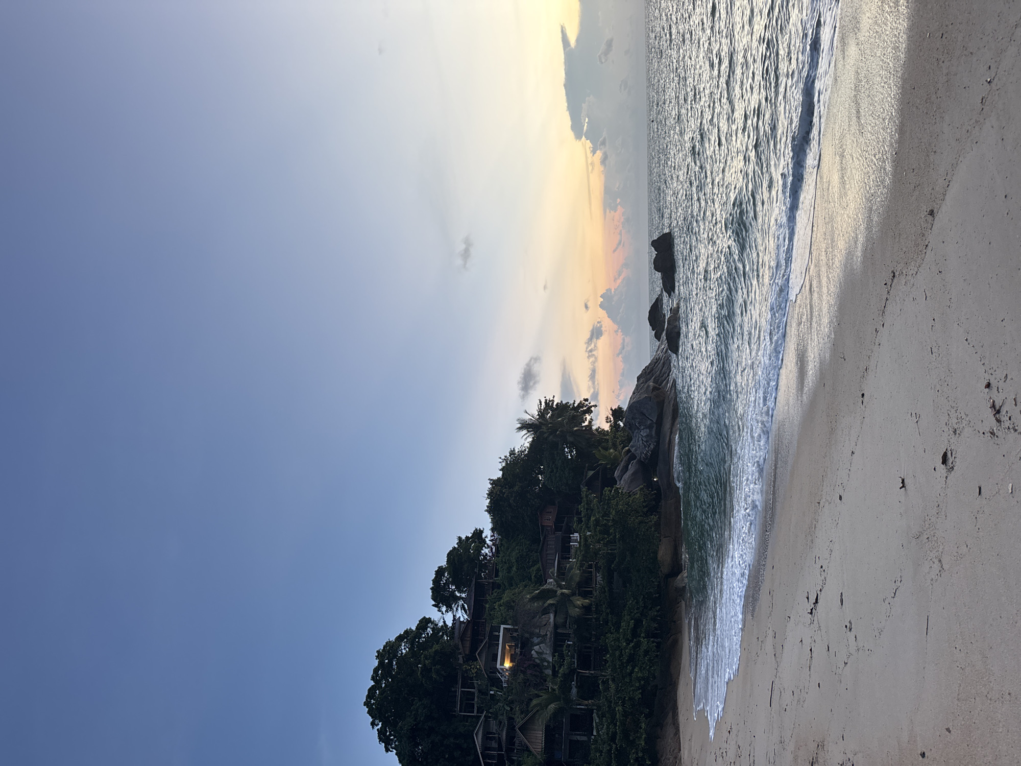 The image depicts a beach scene in Baan Thong Nang, Thailand. The scene shows a beach with white sand, clear water, and vegetation.
On the left side of the image, there is a hillside covered with trees. Several buildings are nestled among the trees on the hillside.
The sky is partly cloudy, with a gradient of blue transitioning to orange at the horizon, indicating sunrise or sunset. The water is calm, with small waves gently lapping on the shore.
The overall scene is peaceful and serene.