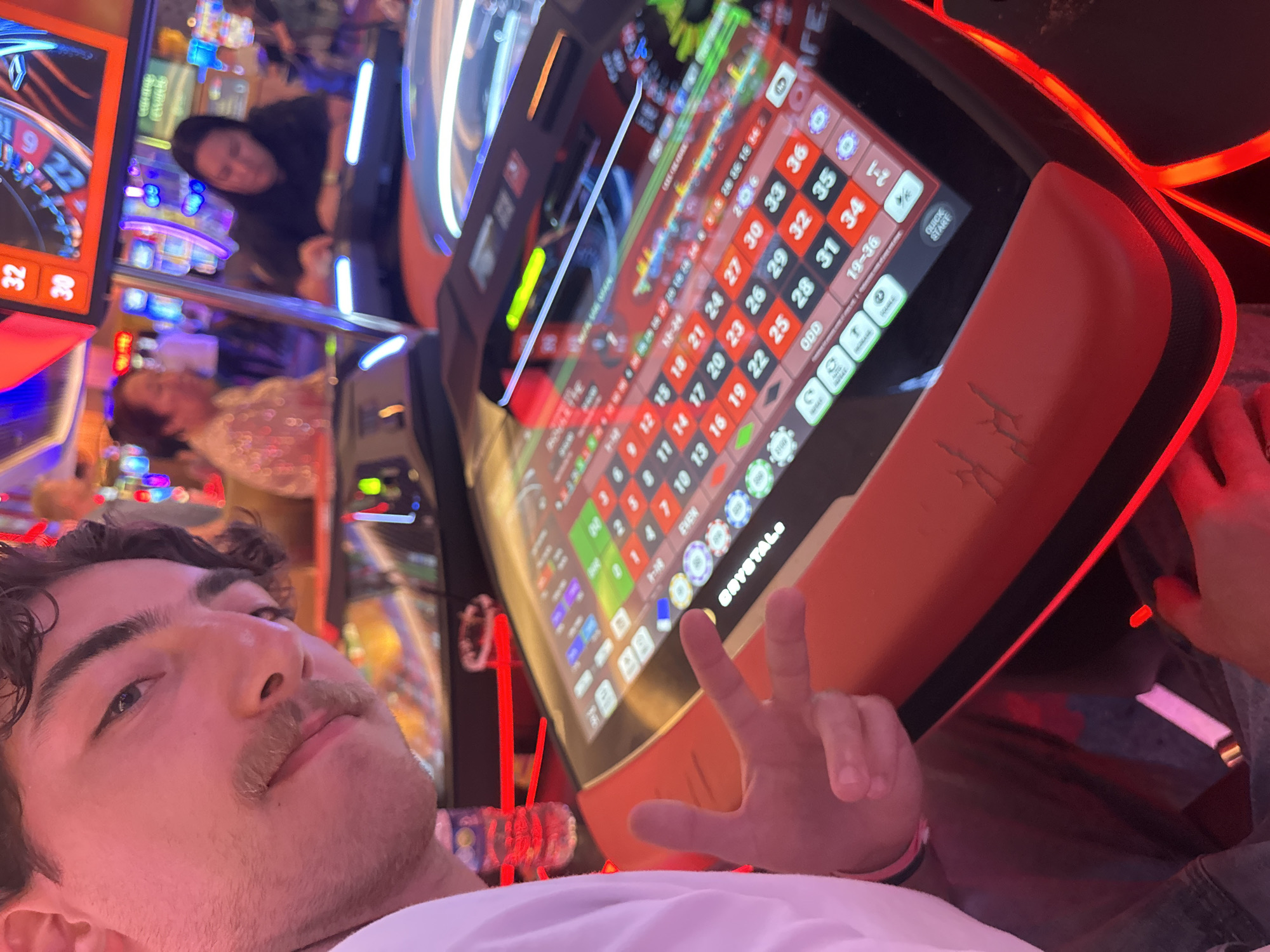 Here is a concise description of the image:
The image appears to be a selfie taken by a person with dark hair and a mustache, posing at a digital roulette table. The table is orange with a black trim and has a screen displaying the roulette layout and betting options. The screen displays the numbers 1-36, as well as 0, 00, and various betting options like "Even", "Odd", "1-2", "19-36". In the background, other casino patrons and gaming machines are visible, suggesting that the scene is in a casino. The logo "CRYSTALE" is visible on the table. The person in the foreground is making a peace sign.
Given the presence of roulette machines and casino environment, it's likely this photo was taken at the Seminole Hard Rock Hotel & Casino in Hollywood, Seminole Reservation, United States of America.