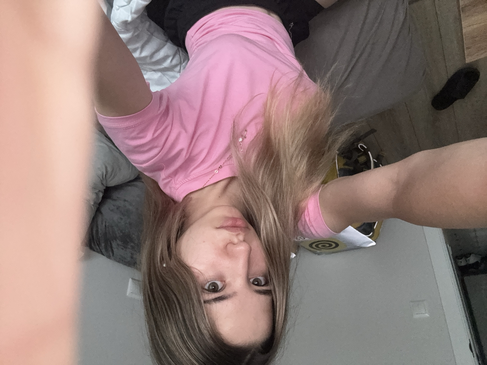 A young woman with blonde hair is taking a selfie in what appears to be a bedroom. She is wearing a pink t-shirt and black shorts. Her left arm is extended, holding the camera, and her right arm is blurred out of the frame. The background includes a bed with gray and white bedding, a wall with a light switch, and a black slipper on the floor. A portion of a box with a spiral design is visible on the bed. The image does not provide any information that would indicate the location as Kazan, Russia.