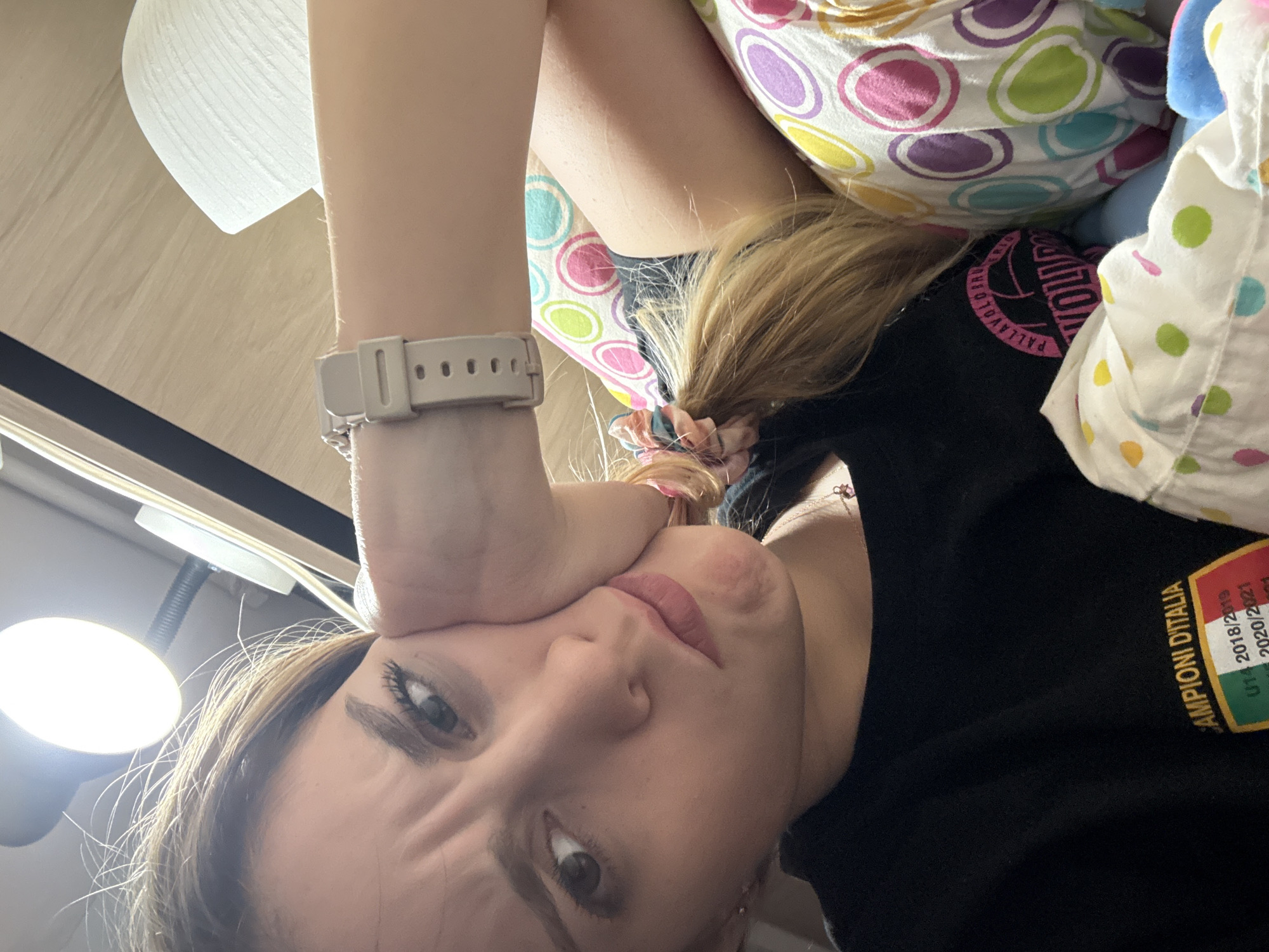A close-up, angled selfie shows a young woman indoors. She has light skin, light brown hair pulled back with a scrunchie, and is wearing a light-colored digital watch on her left wrist. Her left hand is pressed against her face, distorting her expression.
She is lying down, possibly on a bed, with a colorful polka-dot pillow or bedding behind her. She is wearing a black shirt with yellow text and a patch in the shape of the Italian flag, indicating it's a sports uniform. A lamp is visible in the lower left corner of the frame.
The location is unknown, and there is no evidence to suggest it is in Sofia, Bulgaria.