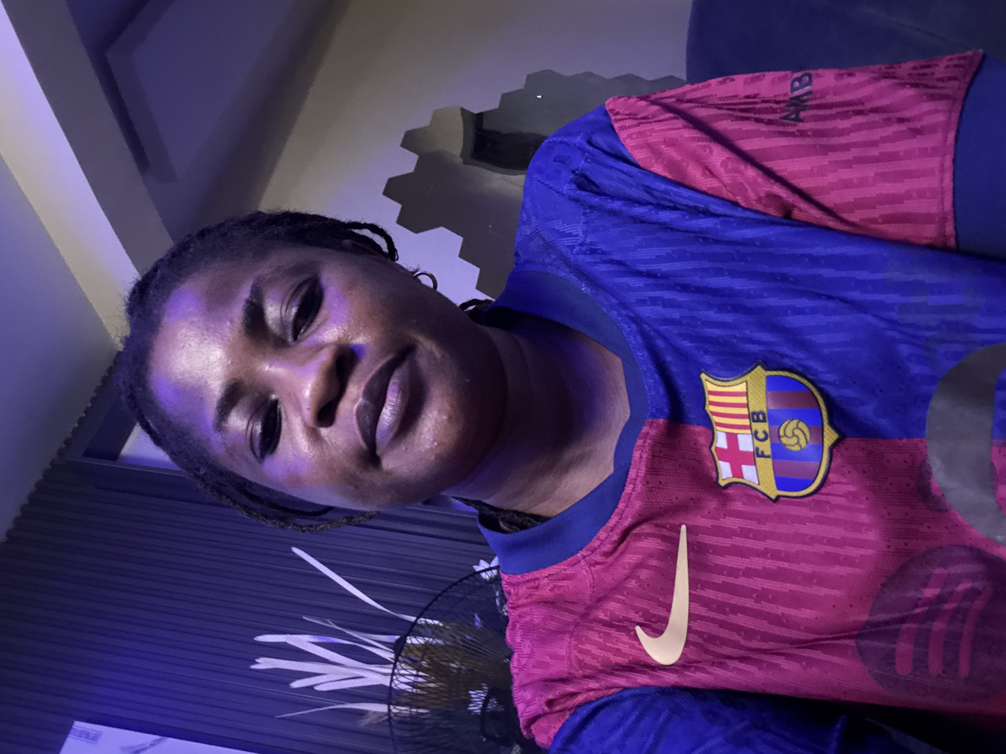 In Ajara, Nigeria, a person with dark skin, braided hair, and a light-colored hoop earring on their left ear is pictured in a close-up, upper-body shot. They are wearing a Barcelona soccer jersey, split vertically between blue and magenta, with the Nike logo on the left chest and the FC Barcelona crest on the right. The person is looking downwards with a neutral expression. The background features a wall with vertical grooved paneling illuminated by purple lighting, and a fan with white decorative elements is visible to the left. To the right, a grey wall displays a geometric, honeycomb-like pattern. The overall lighting is dim with strong blue and purple hues.
