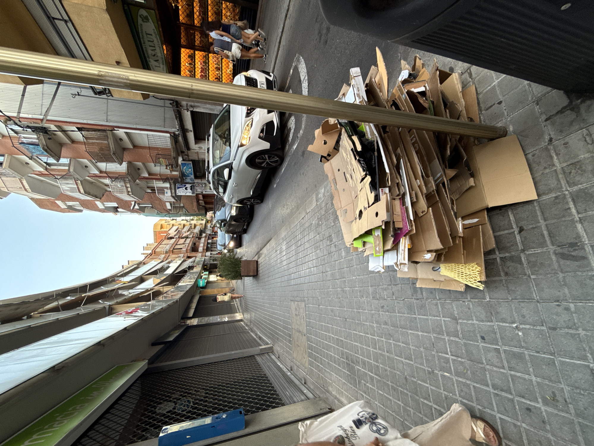 The image captures a street scene in Blanes, Spain, focusing on a pile of flattened cardboard boxes and packaging materials accumulating on the sidewalk in front of a street lamp post. The boxes are of various sizes and are haphazardly stacked, with some showing green markings. To the left of the pile, a section of a building facade is visible, featuring closed storefronts with metal security shutters. A blue electrical box is mounted on the wall. Further down the sidewalk, a person in light-colored clothing and sandals walks away from the camera.
Across the street, another building facade is prominent, with multiple balconies. A white SUV is parked on the street, with its headlights on. Behind the SUV, two individuals are walking towards the right side of the frame. The street is paved with grey tiles, and a yellow mesh protective netting is partially visible within the pile of cardboard. The sky above is clear and blue. The overall impression is that of a commercial street with some accumulated waste.