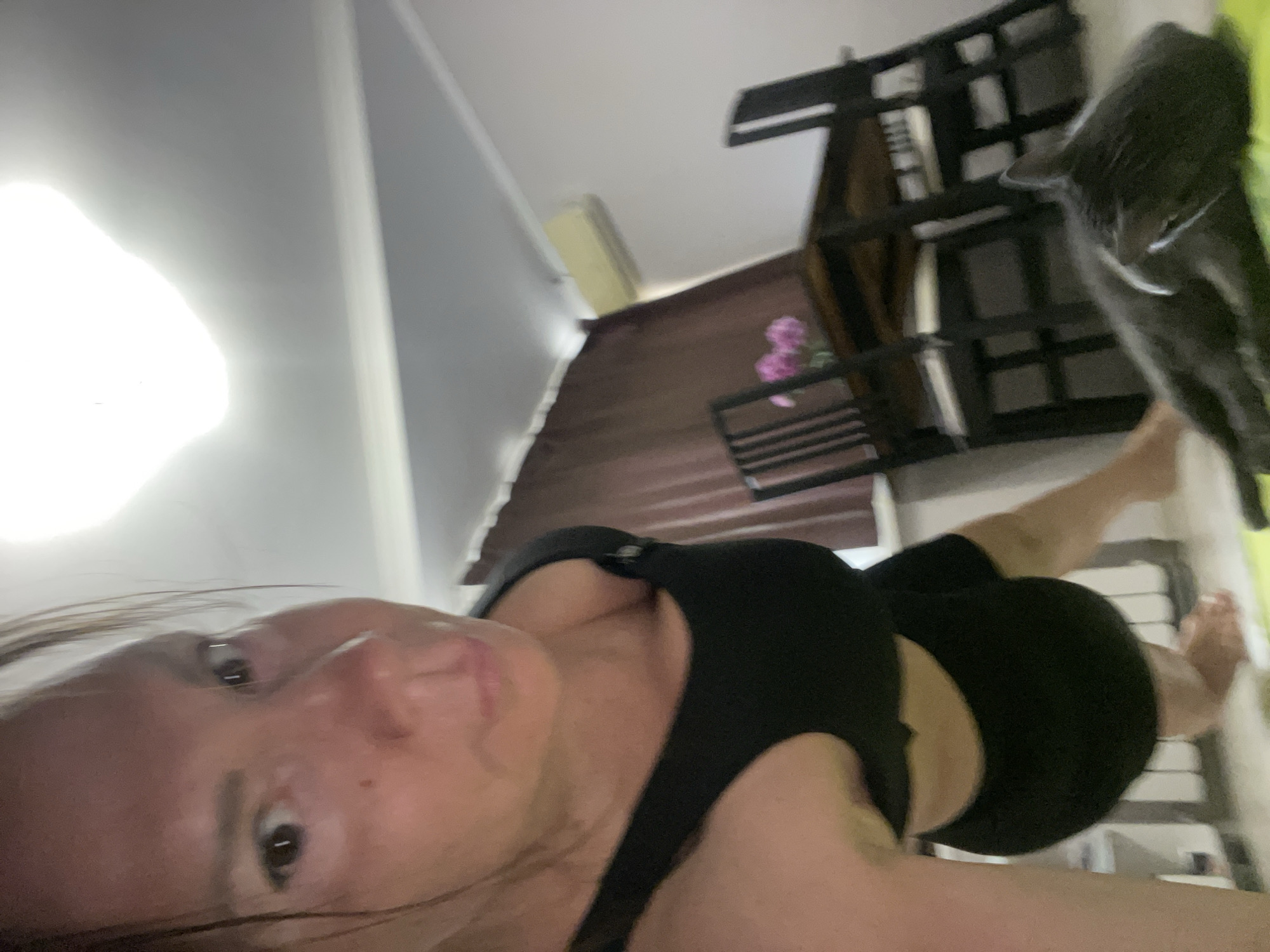 The image captures a low-angle, close-up view of a woman in athletic wear, appearing to be in a room that might be a home gym or living space. Her face is prominent in the foreground, with sweat visible on her skin, suggesting recent physical activity. She is looking directly at the camera. Behind her, a dining table with chairs is visible, along with a curtain and an air conditioning unit on the wall. A gray cat is lying on a green yoga mat in front of the table and chairs. The lighting suggests an indoor environment, possibly with artificial light from above. The overall impression is a candid shot taken during or after a workout. There is no information or visual cue within the image that would allow for identification of the specific location within the city of Sochi, Russia.