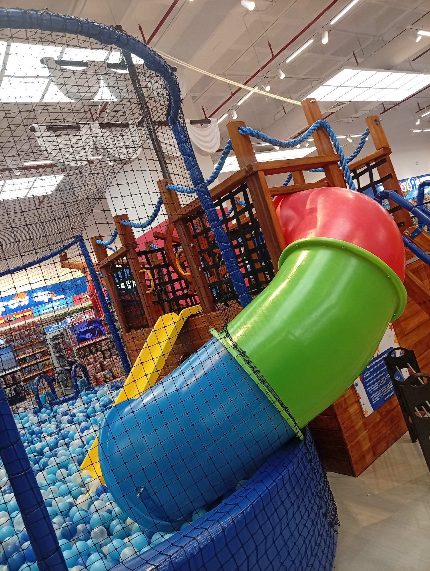 The image depicts an indoor playground in Porlamar, Venezuela. The playground features a ball pit filled with blue and white balls, a yellow slide, a red, green, and blue tube slide, and a wooden climbing structure with blue rope accents. The playground is enclosed by netting. In the background, shelves of merchandise are visible, indicating the playground is located within a retail store. The lighting is bright, and the overall environment appears clean and well-maintained. No people are visible in the frame.