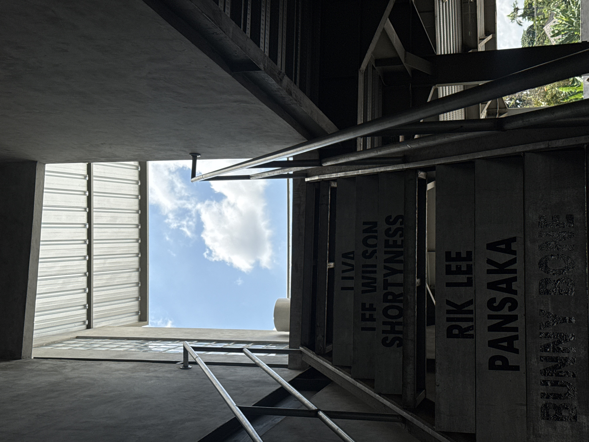 This low-angle shot depicts a structure with concrete and metal elements, possibly a building or open-air construction in Mengwi, Indonesia. The perspective is upward, framing a patch of blue sky with clouds. Staircase steps are visible, with names printed vertically on each step: "LIVA," "IFF WILSON," "SHORTYNESS," "RIK LEE," "PANSAKA," and "BUNNY BONE." Metal railings accompany the staircase. Architectural details include corrugated metal roofing and concrete beams. Green foliage is glimpsed in the upper right corner, indicating outdoor vegetation.